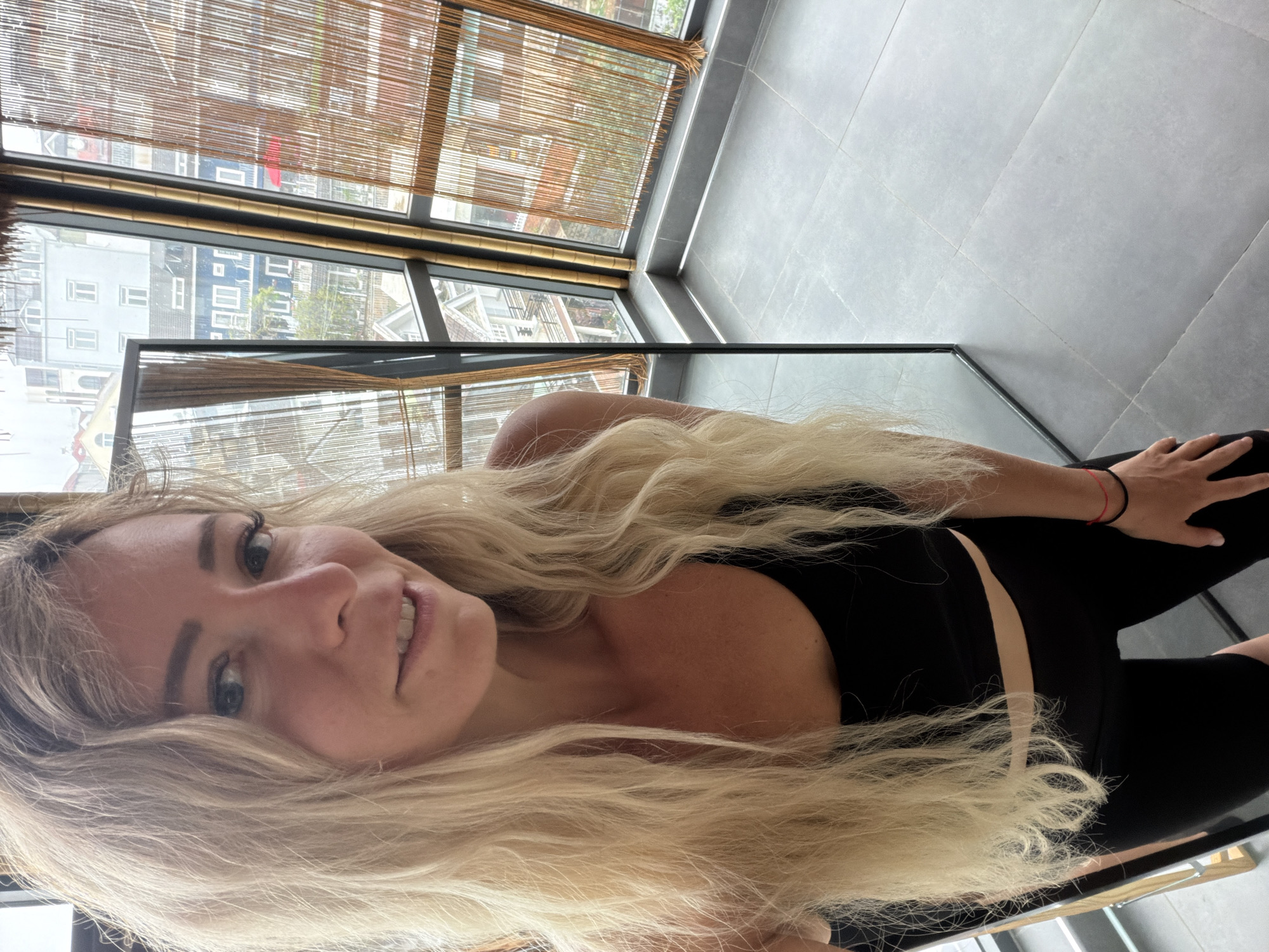 A woman with long blonde hair and fair skin is taking a selfie in an indoor setting. She is wearing a black sports bra and black leggings. Her right hand is visible, wearing a red and black bracelet. Behind her, a large mirror reflects her and the surrounding environment. The background shows large windows with bamboo blinds, offering a view of buildings and a street, suggesting an urban setting. The floor is tiled in a grey, stone-like pattern. The architecture visible through the windows appears to be in the style of Da Lat, Vietnam, with multi-story buildings and a visible sloped roof, consistent with the city's French colonial and modern influences. The lighting suggests an overcast day.