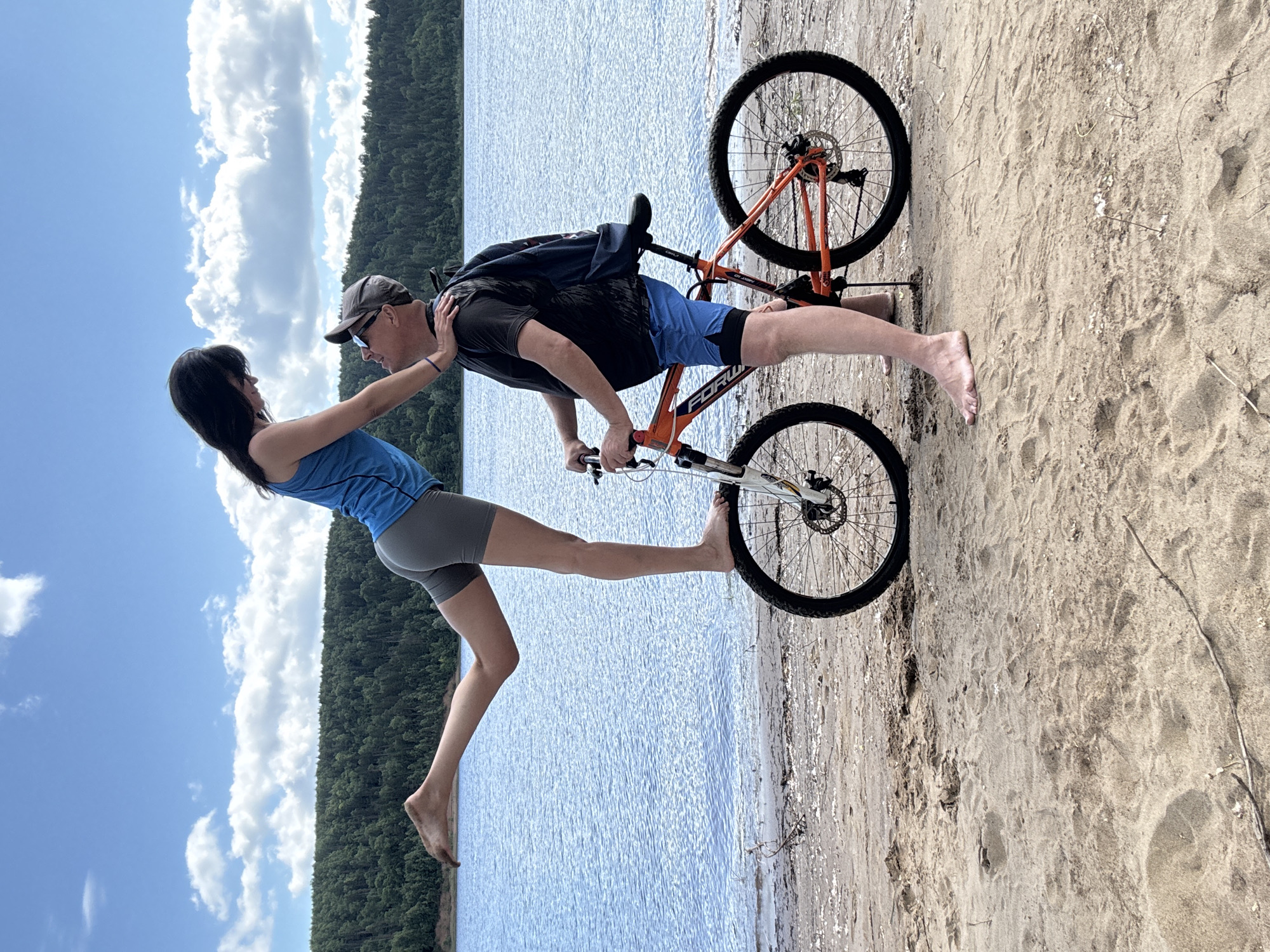 A woman with dark hair, wearing a blue tank top and gray shorts, is balancing on the front tire of an orange and black bicycle. Her left leg is extended backward. She is looking down at a man who is standing next to the bicycle, holding the handlebars. The man is wearing a gray baseball cap, sunglasses, a black t-shirt, and blue shorts. He has a dark backpack on. Both individuals are barefoot. They are on a sandy beach next to a large body of water. In the background, there is a dense forest lining the opposite shore. The sky above is blue with scattered white clouds. The image appears to have been taken on a sunny day. The overall scene suggests a recreational activity by a lake.