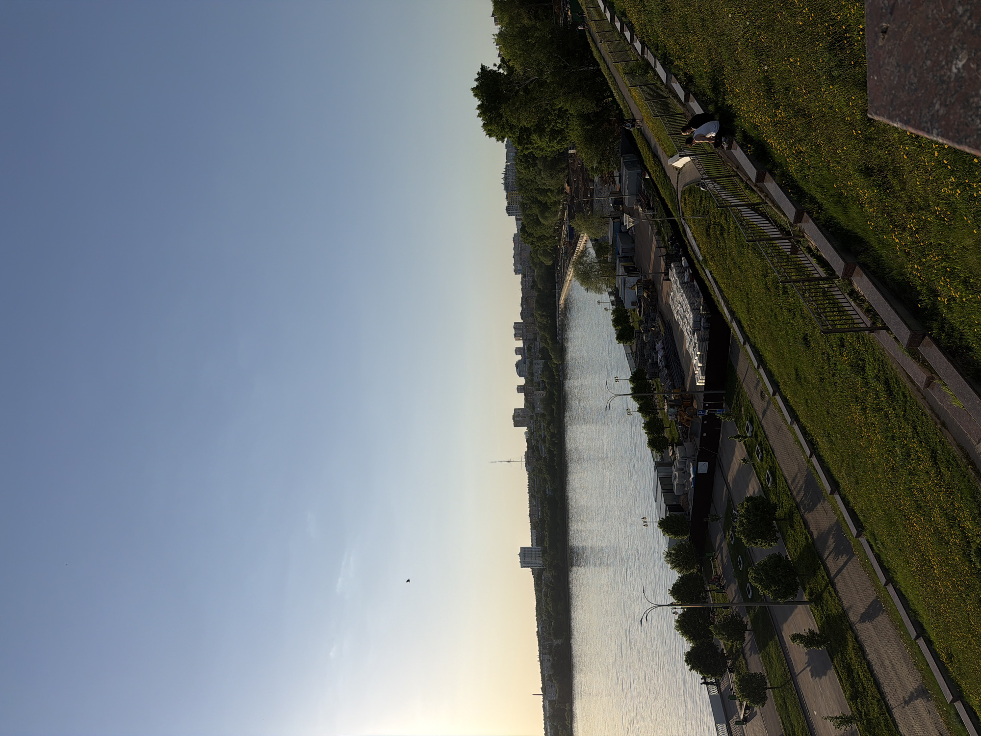 View of Izhevsk, Russia, overlooking the city's embankment, a body of water, buildings, trees, sidewalks, and grassy areas. Two people are visible near a fence on the upper-right side. A bird flies across the sky. The sky transitions from a bright blue to a soft yellow near the horizon.