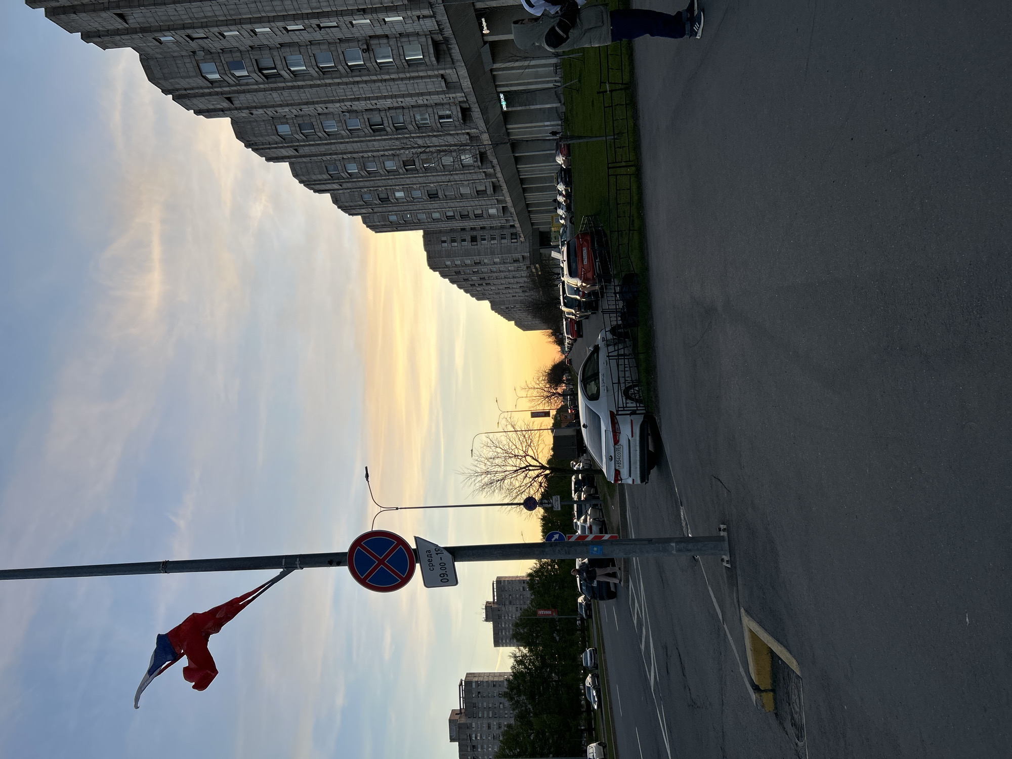 The image shows a street scene in Saint Petersburg, Russia, at sunset. A tall, gray, Soviet-era apartment building dominates the upper left of the frame. The sky is a gradient of blue to orange. Along the street, cars are parked next to a small strip of grass. A traffic sign indicating "No Stopping" in red and blue is visible on a pole, with a Russian flag also attached. In the background, more apartment buildings are visible. A person is visible in the upper right. The foreground consists primarily of the asphalt street surface.