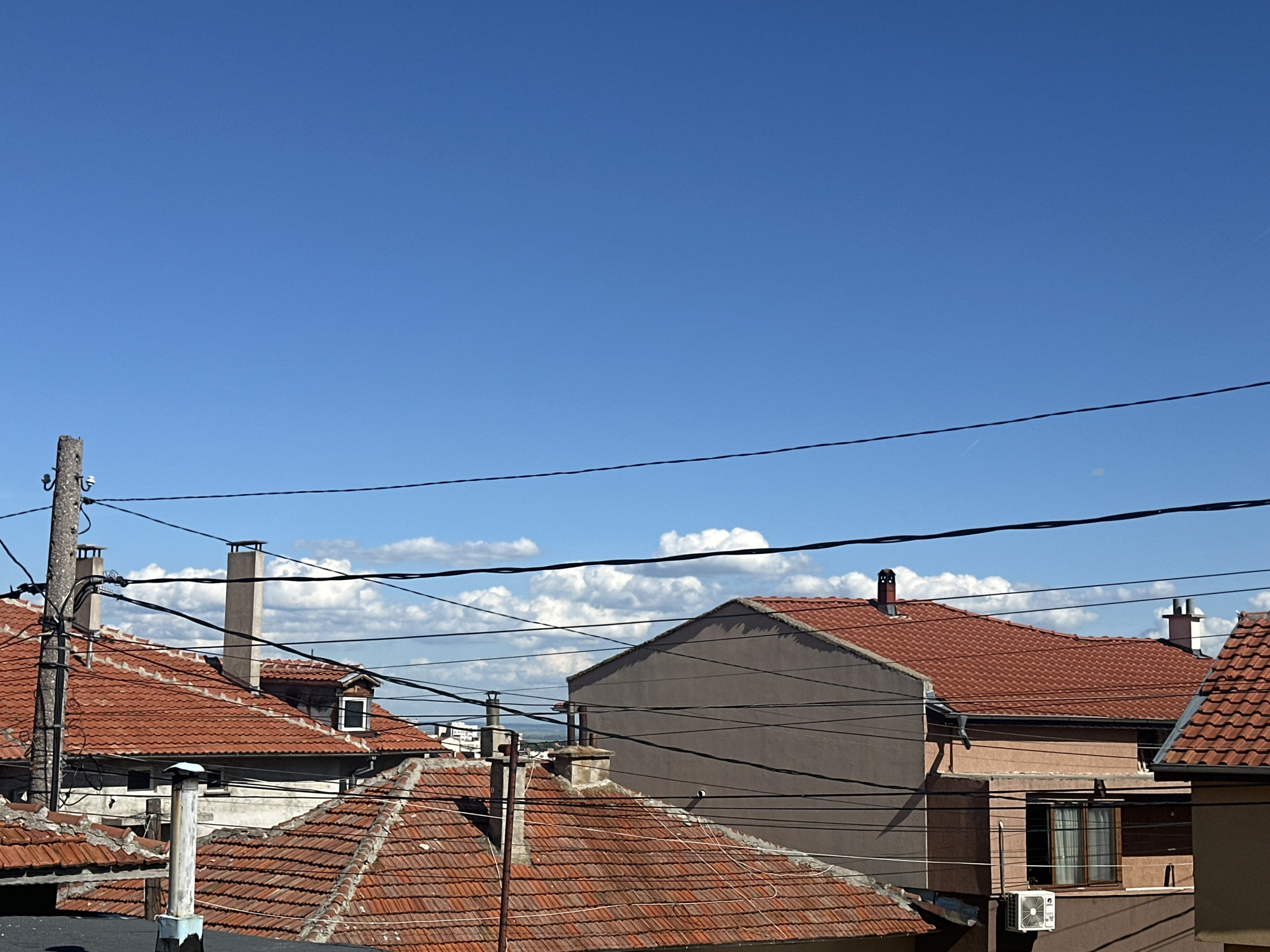 The image shows a daytime view of rooftops in Sliven, Bulgaria. The rooftops are covered in red clay tiles. Power lines cross the view. There is a mix of residential structures with chimneys and varying heights. A utility pole is seen on the left side of the image. The sky is blue with scattered white clouds.