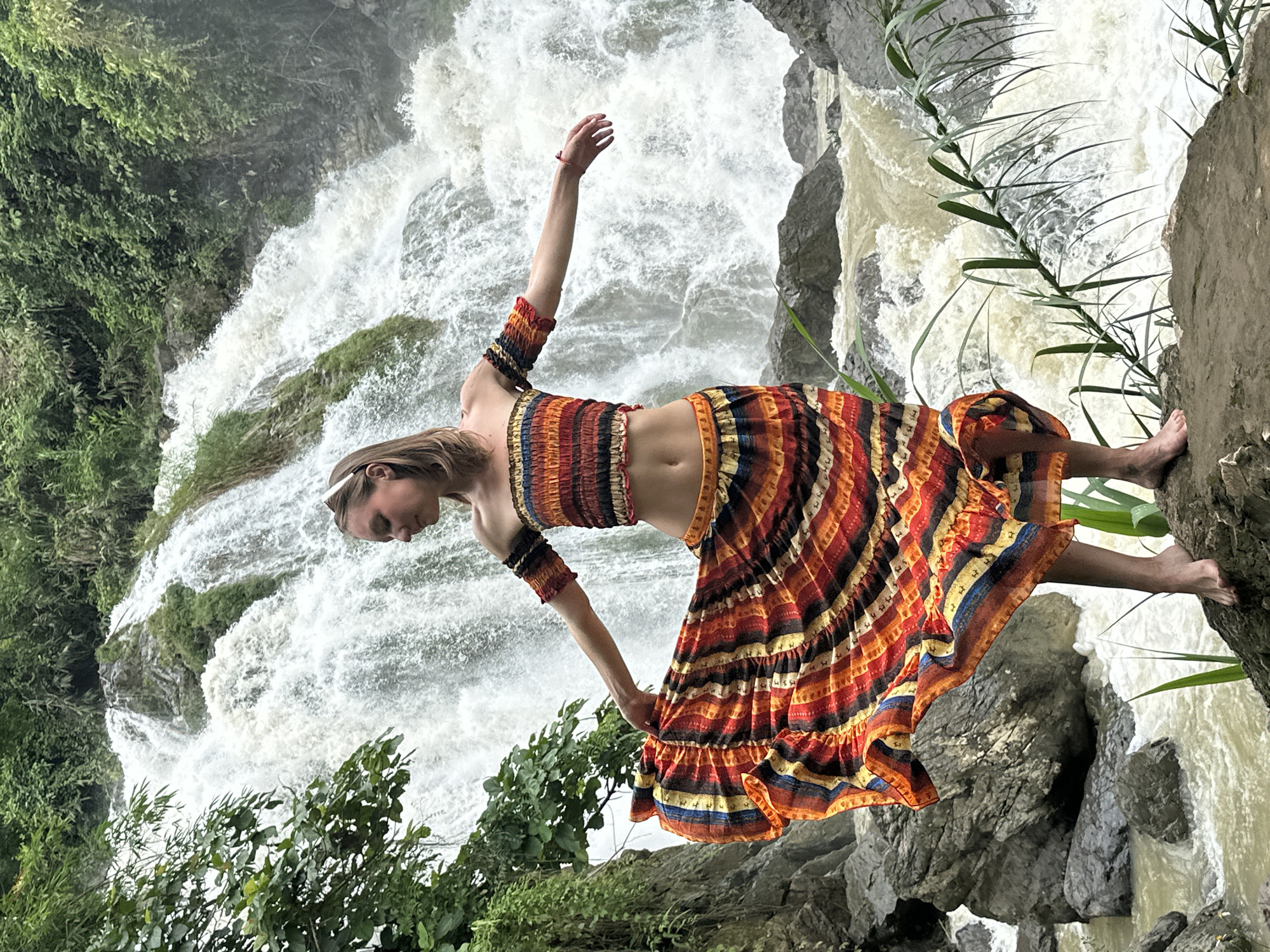 A woman stands on a rock in front of a cascading waterfall in Bản Phúc, Vietnam. She is wearing a colorful off-the-shoulder crop top and a matching flowing skirt. Her arms are outstretched as if dancing or posing. The waterfall is wide and powerful, with lush green foliage surrounding it. The ground where she stands is rocky, and there is a river flowing below.