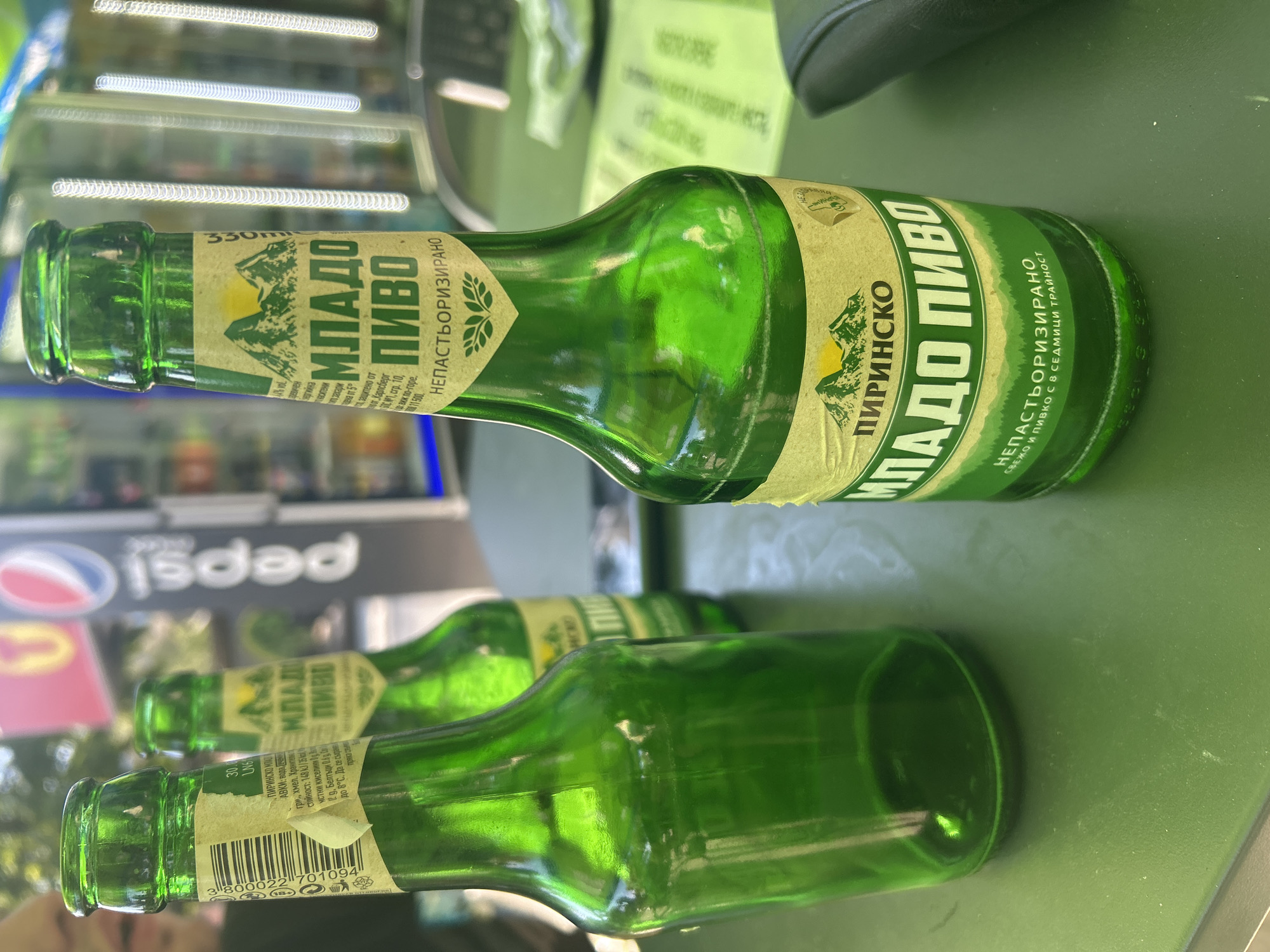 Two green glass beer bottles of "Pirinsko Mlado Pivo" are on a green surface, likely a table. One bottle is in focus in the foreground, displaying its label with mountains and text. Another bottle is behind it, slightly out of focus. A third, mostly empty, bottle is to the left. In the background, a Pepsi logo is visible on a dark sign, along with blurred figures and foliage, suggesting an outdoor or semi-outdoor setting, possibly a beer garden or cafe in Sofia, Bulgaria.
