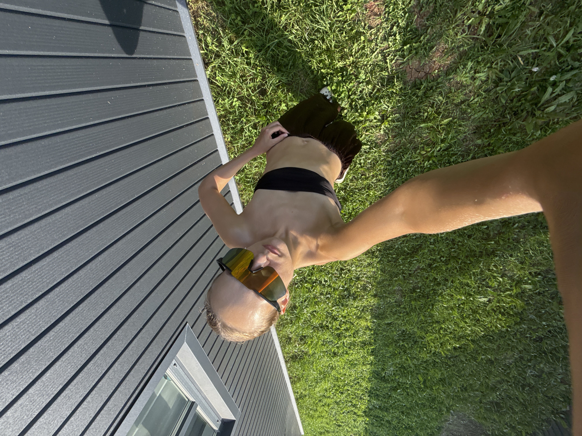 The image is a high-angle, full-body shot of a young person outdoors, standing on a grassy area next to a dark, vertically-paneled building. The person is wearing large, mirrored sunglasses with orange lenses, a black bandeau top, and dark, possibly brown, patterned shorts. Their left arm is extended towards the camera, holding it, indicating a selfie. The sun is casting a shadow of the building onto the grass. The building appears to be a modern structure with a window visible on the left side. The overall scene suggests a warm, sunny day. There is no identifiable information in the image that places it specifically in the city of Подшивалово, Russia.