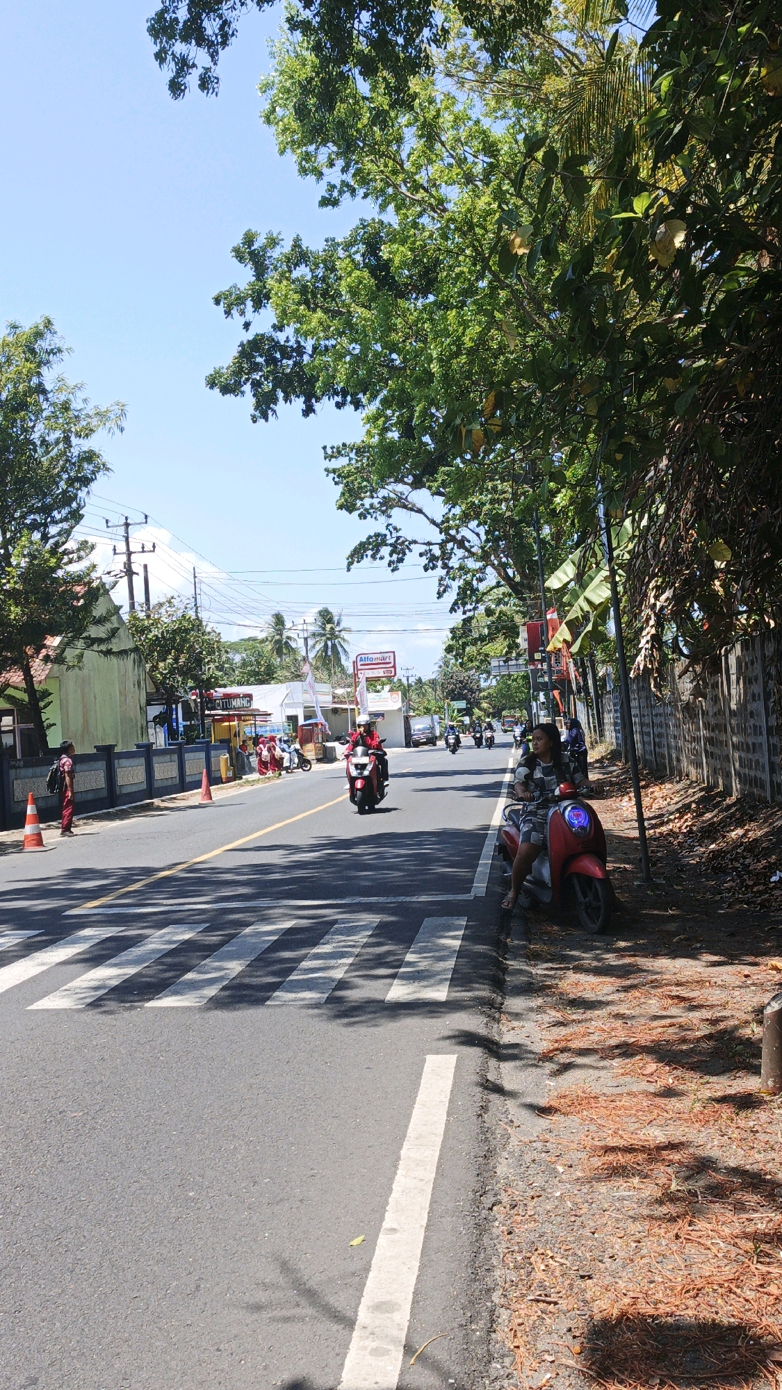 The image shows a street scene in Cibenda, Indonesia. A paved road runs horizontally across the frame, with a pedestrian crossing marked by white stripes. On the right side of the road, a red scooter is parked next to a person sitting on it, wearing a patterned shirt and shorts. A concrete wall runs along the right edge of the road, with trees and foliage overhead, casting shadows on the ground. Fallen orange leaves are scattered on the shoulder of the road.
On the left side of the road, several motorbikes are in motion, including one red scooter with a rider wearing a red jacket and helmet. A person in red pants and a backpack stands near a traffic cone on the sidewalk. Further down the road, buildings are visible, including one with a green facade and a sign for "Alfamart." Other buildings appear to be shops or residences. The sky is clear and blue, indicating a sunny day. The overall scene depicts a typical street in a residential or commercial area of Cibenda.