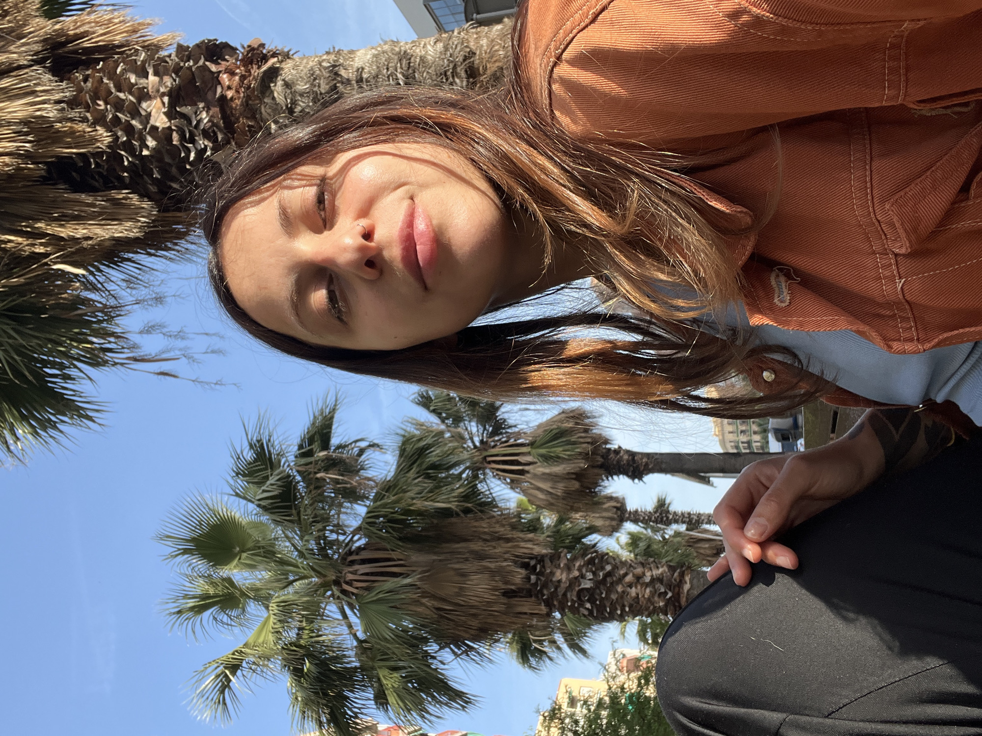 A woman with brown hair and an orange jacket is in a park in Barcelona, Spain. She has dark eyes and a nose piercing. She is sitting down wearing black pants and has a tattoo on her left wrist. Palm trees are visible in the background, as well as a clear blue sky. In the distance, buildings and trees are seen.