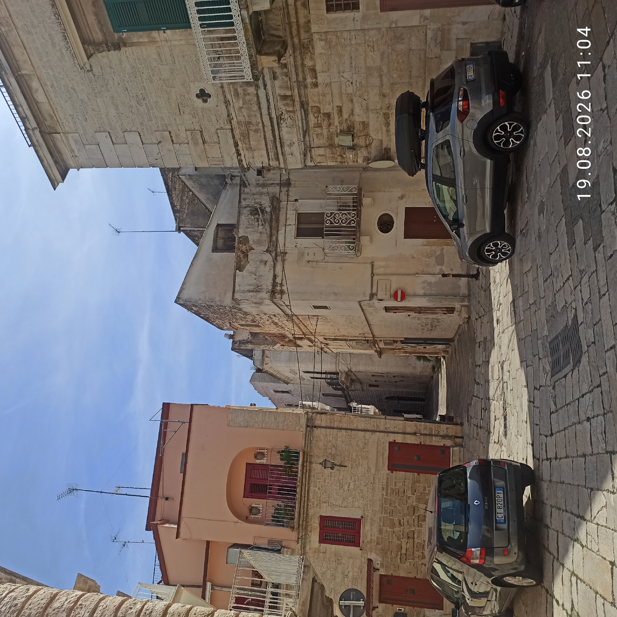 The image shows a narrow, cobblestone street in Ruvo di Puglia, Italy, lined with historic stone buildings. The buildings feature traditional architecture with arched doorways, red shutters, and balconies with decorative ironwork. A light-colored car, a grey SUV with a roof box, is parked on the right side of the street, and a dark-colored hatchback is parked on the left. The sky is clear and blue, with sunlight casting shadows on the cobblestones. A street sign indicating "VIA MADONNA" is visible on the right. The scene captures a typical streetscape in a historic Italian town.
