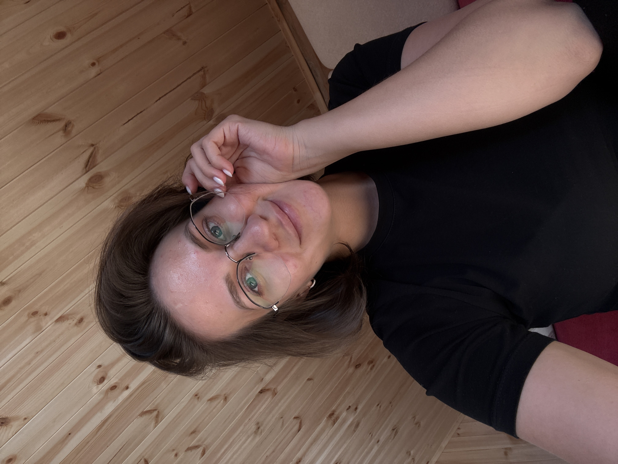 A headshot-style selfie captures an adult female positioned indoors in what is identified as Lenino, Russia. She has brown, shoulder-length hair and wears wire-rimmed glasses with a silver-colored frame, through which her green eyes are visible. Her expression is a slight smile, and her right hand, with white-painted fingernails, is raised, fingers gently touching the side of her glasses near the temple. She is dressed in a plain black short-sleeved t-shirt. The background consists of light-toned wooden paneling, arranged diagonally, indicative of a sloped ceiling or wall. A small portion of a reddish upholstered surface is visible in the lower right corner of the frame.