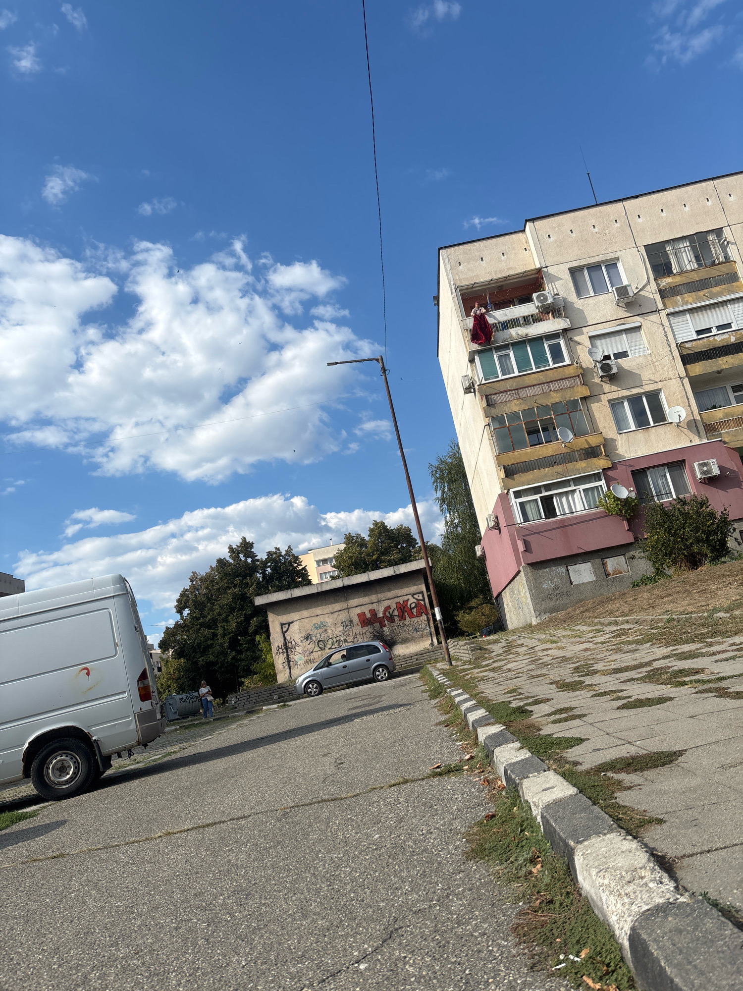 The image captures an outdoor scene, likely in Blagoevgrad, Bulgaria, on a sunny day with scattered clouds. The perspective is tilted, giving a dynamic feel to the urban environment.
In the foreground, a white van is parked on an asphalt road. The road has distinct cracks and is bordered by a curb made of alternating black and white concrete blocks, with patches of grass growing between them.
Dominating the midground and background is a multi-story apartment building with a light-colored facade, adorned with numerous balconies, windows, and air conditioning units. On one of the upper balconies, a person is visible, hanging a bright red piece of fabric, possibly clothing or a blanket. Another, smaller building with graffiti is situated in front of the apartment block. A silver car is parked near this smaller building, and a person in blue jeans and a light-colored shirt stands beside it, near a dumpster. Trees with lush green foliage are interspersed between the buildings. A tall streetlamp stands near the apartment building.
The sky is a vibrant blue, with fluffy white clouds drifting by, indicating fair weather. The sunlight suggests it is daytime, likely late morning or early afternoon. The overall mood of the image is one of everyday urban life, with a hint of candidness due to the person hanging laundry on the balcony.
Visible text includes graffiti on the smaller building, with some letters and the numbers "2025" discernible.