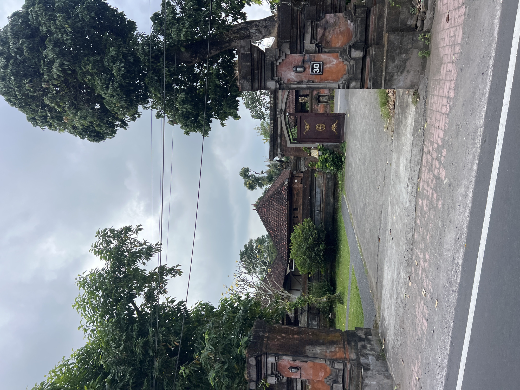 The image shows an ornate gateway in Abiansemal, Indonesia, leading into a traditional Balinese compound. The gateway is made of weathered stone and brick, with intricate carvings and decorative elements. The gate itself is a dark brown metal with gold detailing and is partially open. Above the gateway, a sign reads "GERIA SADA."
To the left and right of the gateway are large, mature trees with dense green foliage. The sky above is overcast and cloudy, suggesting a potentially rainy day. Beyond the gateway, a paved driveway leads to traditional Balinese houses with distinctive red tile roofs. Green grass and lush vegetation are visible within the compound. A number "30" is displayed on a black plaque mounted on the right gatepost. The foreground shows a paved street with a white line marking the edge, indicating the location is along a public road. The overall impression is one of a serene and culturally rich entrance to a private residence.