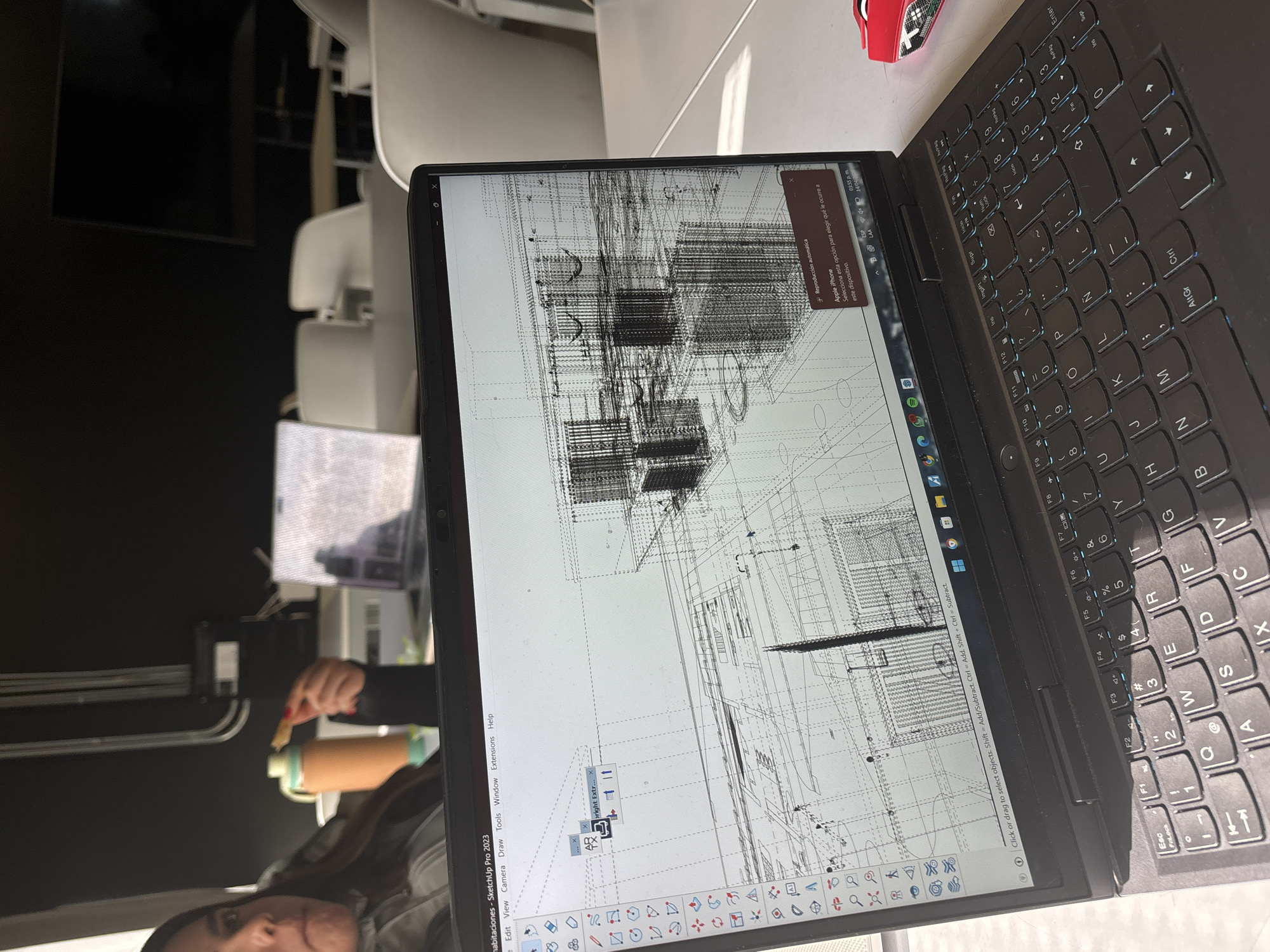 Here is a matter-of-fact description of the image:
The image shows a close-up of a black laptop with a design software interface (SketchUp Pro 2023) on the screen displaying a building plan. The laptop is on a light-colored surface, possibly a desk, next to a small red object resembling a car. The laptop keyboard is visible. A person is visible in the lower-left corner holding a brown object. In the background, there are chairs and what appears to be another electronic device and a black wall. The location cannot be determined based on this image.