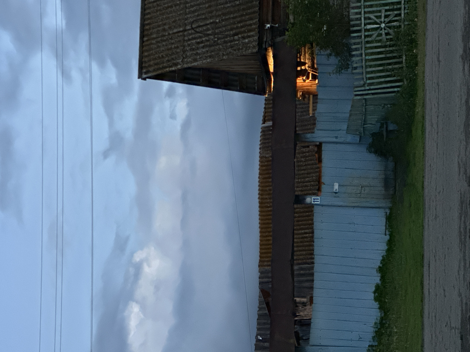 A close-up, eye-level outdoor shot captures a dimly lit rural scene at dusk. The sky is a blend of dusky blue and grey, with dark clouds suggesting impending weather. Four thin power lines stretch horizontally across the upper portion of the frame.
In the mid-ground, a dilapidated wooden fence, painted a faded blue, runs horizontally across the image. To the right of the fence, a section of a weathered, corrugated roof is visible, supported by dark wooden beams. A warm orange light emanates from behind this roof, hinting at an interior light source. The fence has a simple, weathered wooden gate with a rectangular sign above it showing the number "11". To the right of the blue fence, a section of a green, slatted wooden fence is visible.
The foreground is occupied by a strip of dark, patchy asphalt, with a narrow band of green grass and some sparse vegetation along the base of the fence. The overall impression is one of a quiet, possibly abandoned or sparsely inhabited rural dwelling as evening sets in.
