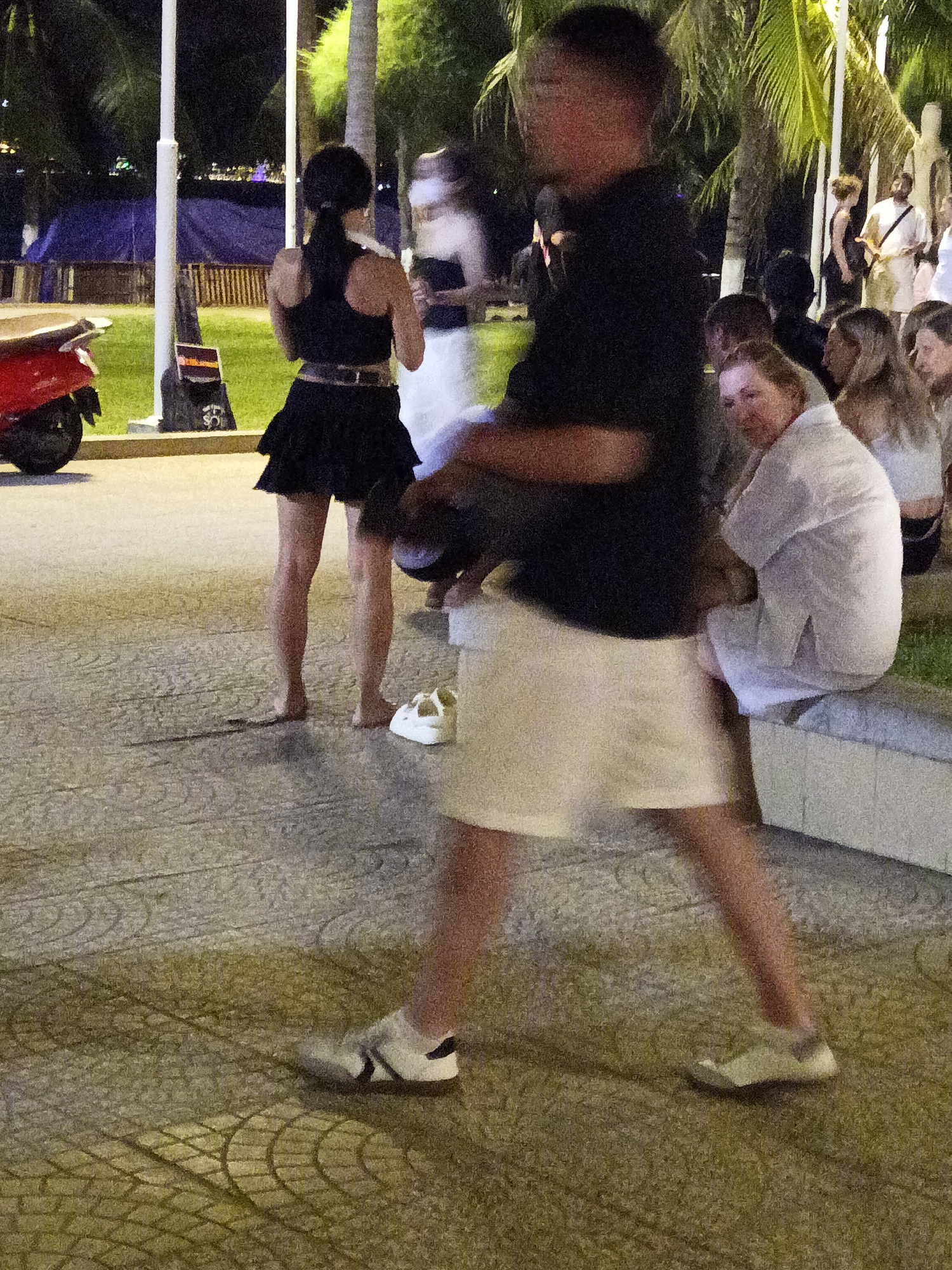 The image depicts a street scene at night in Nha Trang, Vietnam. In the foreground, a person in a black top and white shorts is walking from right to left, their face blurred by motion. To the left, a person in a black top and a short black skirt is standing, facing away from the camera. Behind them, a red scooter is parked. Further back, several other people are visible, some seated on a low wall, with palm trees and foliage forming the background. The ground is paved with textured tiles. The overall ambiance is that of a public space at dusk or night.