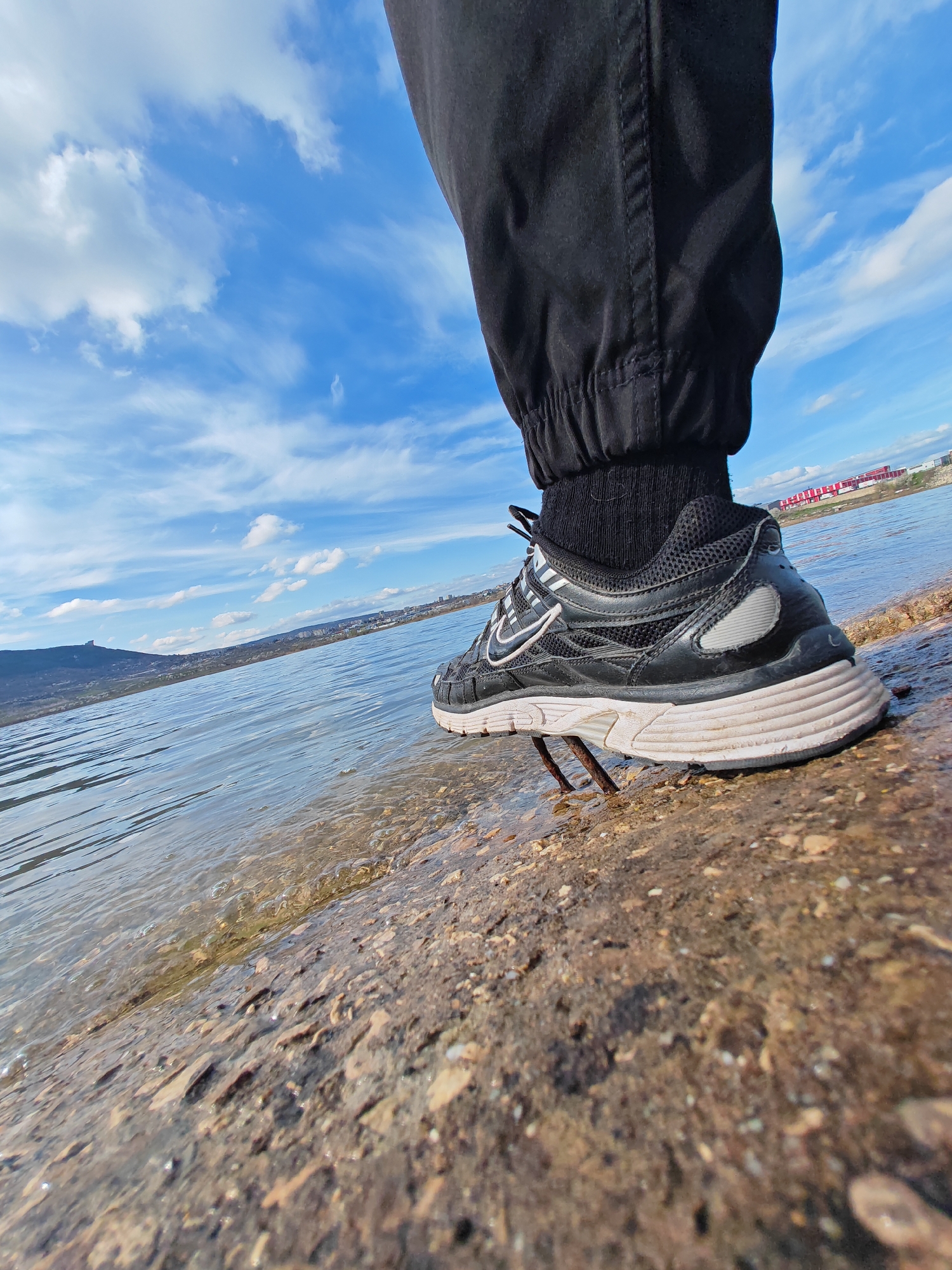 A person's right foot is positioned near the edge of a body of water in a city in Shumen, Bulgaria. The person wears black joggers with a cuffed ankle over a black sock. The black and white Nike shoe is resting on rusted metal rods protruding from the brown rocky ground. The water is clear and gently lapping against the shore. In the background, there is a cityscape with buildings and a mountain. The sky is blue with scattered white clouds.