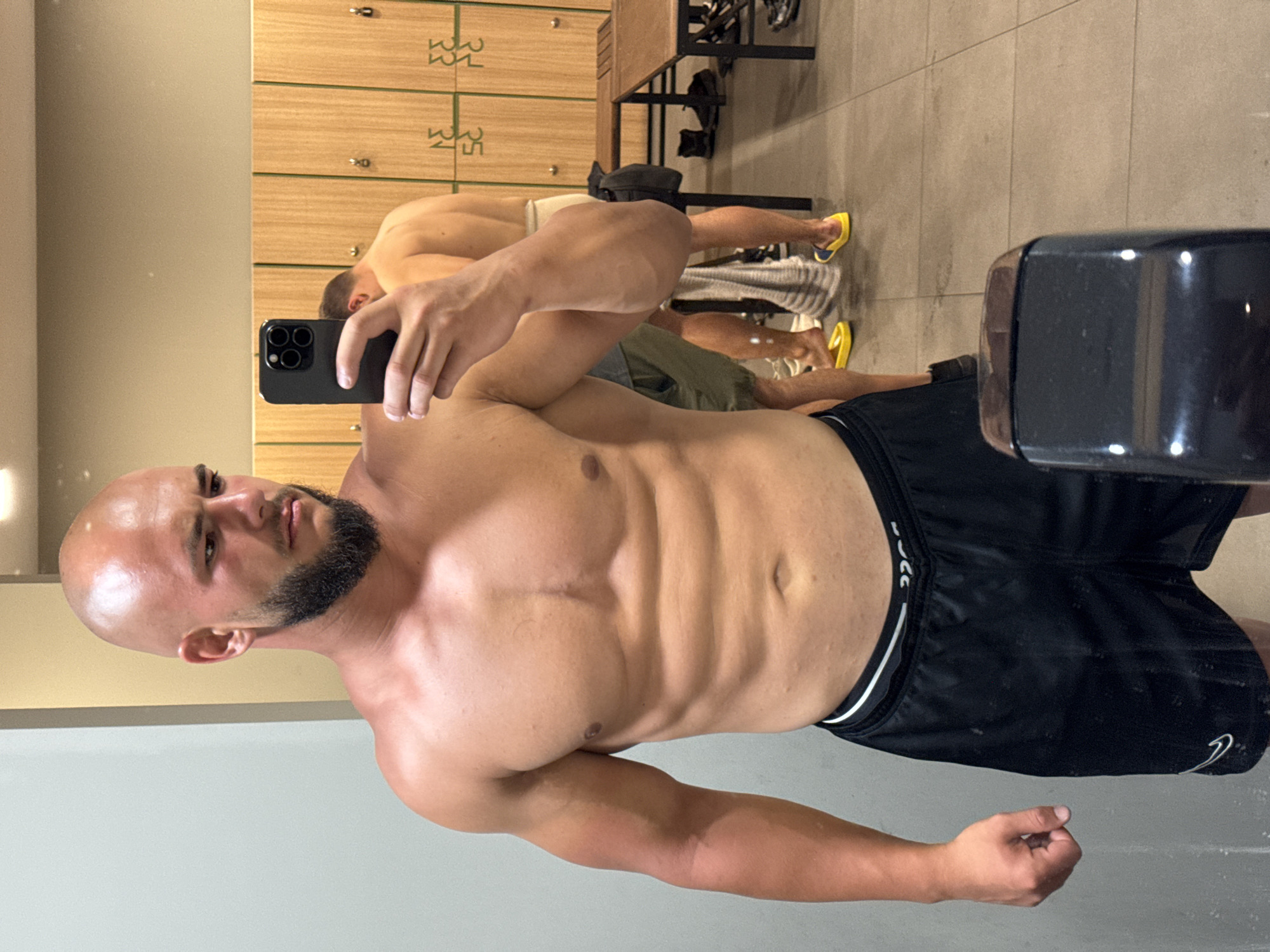 A bald, bearded man with a muscular physique is taking a selfie in a gym locker room. He is wearing black shorts. In the background, another man is visible from the back, and lockers line the wall. There are benches and shoes on the floor. The man is holding a black smartphone. The location is the locker room of a gym in Sofia, Bulgaria.