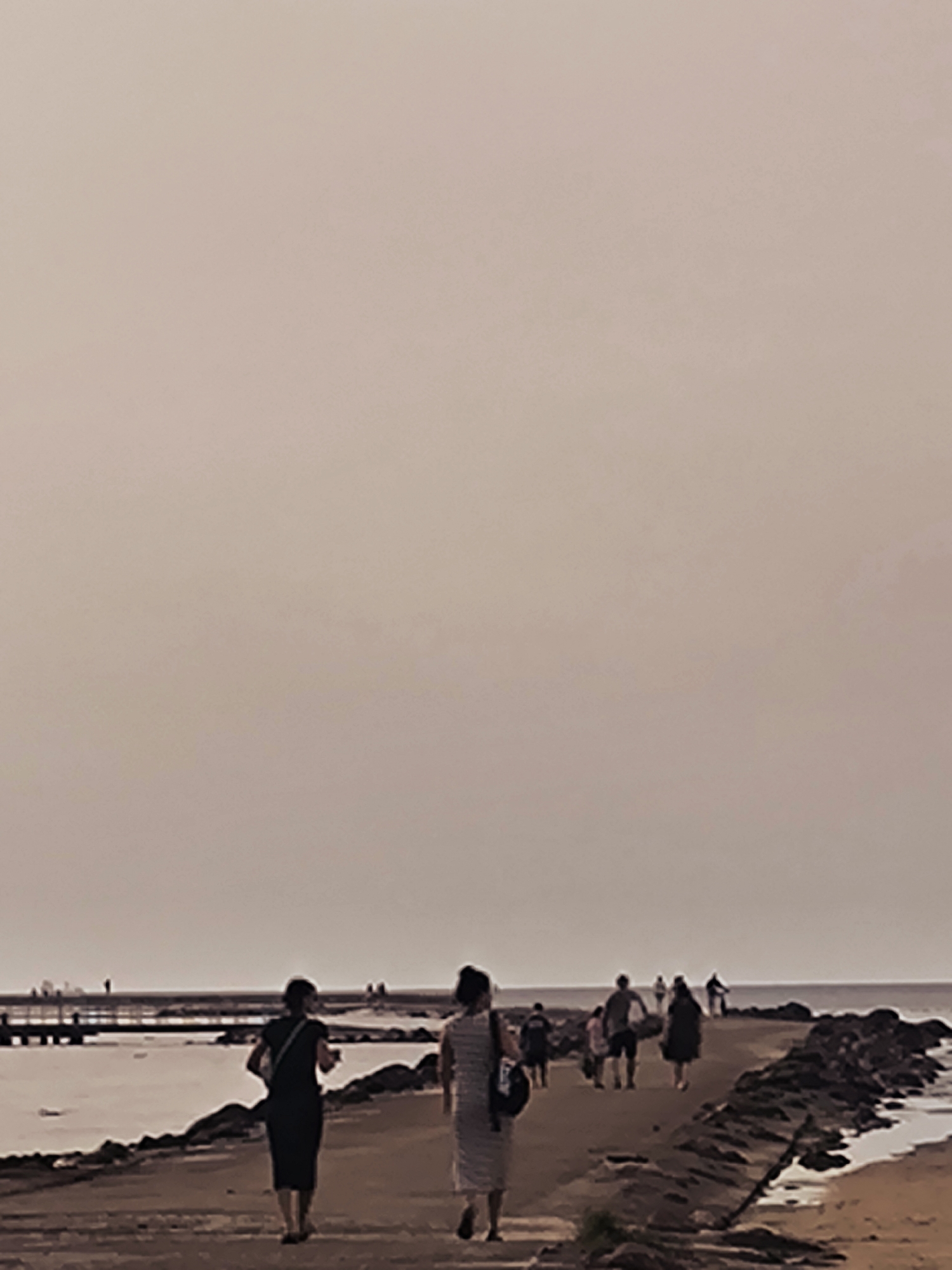 The image shows a view of the Daugava River in Riga, Latvia, on a hazy day. Several people are walking along a pier or embankment. In the foreground, two women are walking away from the camera. The woman on the left wears a dark dress and carries a bag over her shoulder. The woman on the right wears a striped dress. Further down the pier, other figures are visible, some walking in groups. The water of the river is calm, and on the far side, there appears to be a distant structure with some small figures on it. The overall atmosphere is subdued due to the overcast and hazy conditions, with muted colors dominating the scene.