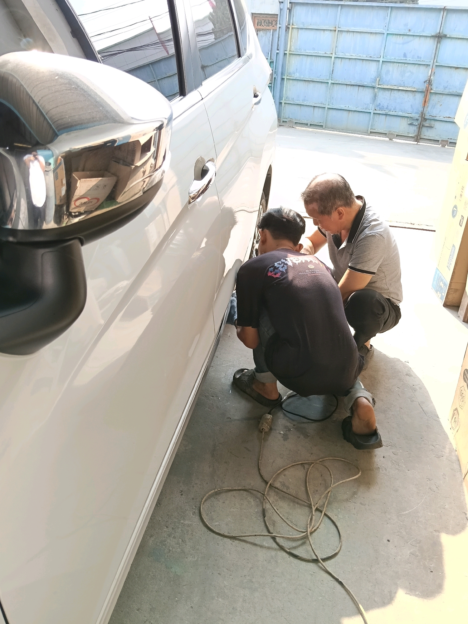 In Belimbing, Indonesia, two men are working on the side of a white vehicle, possibly a car or van. One man, wearing a dark t-shirt with a graphic design and jeans, is crouched down near the vehicle's wheel. He is holding a tool, possibly a sander or polisher, connected to a power cord that lies on the ground. The other man, older and wearing a grey polo shirt and dark pants, is also crouched, observing or assisting the first man. The scene appears to be outdoors, with a concrete floor and a large metal gate or wall in the background. The lighting suggests it is daytime. There are cardboard boxes visible to the right of the frame.