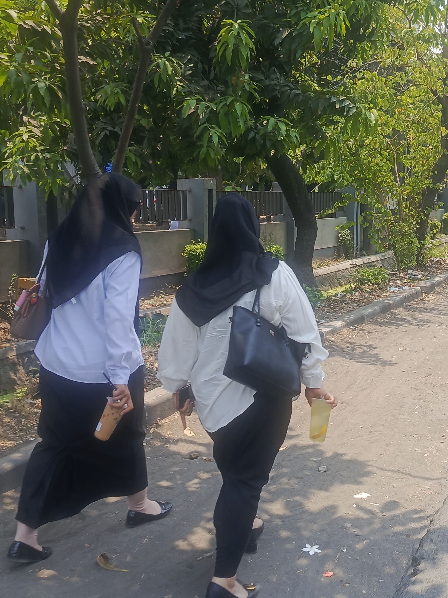 Two women, each wearing a black hijab and a white long-sleeved collared shirt with black pants or skirt, are walking away from the camera on a paved surface. The woman on the left is wearing a black skirt and holding a brown bag over her left shoulder and a clear plastic cup with a brown beverage and a straw in her right hand. The woman on the right is wearing black pants and carrying a black tote bag on her right shoulder and holding a clear plastic cup with a yellow beverage and a slice of lemon in her right hand. They are walking on a sidewalk or path adjacent to a street. In the background, there are trees with green foliage, a grey concrete barrier or wall, and a metal fence. The scene appears to be outdoors in a tropical or subtropical environment, possibly a campus or park, given the presence of trees and the bright sunlight casting shadows. The location is identified as Waru, Indonesia.