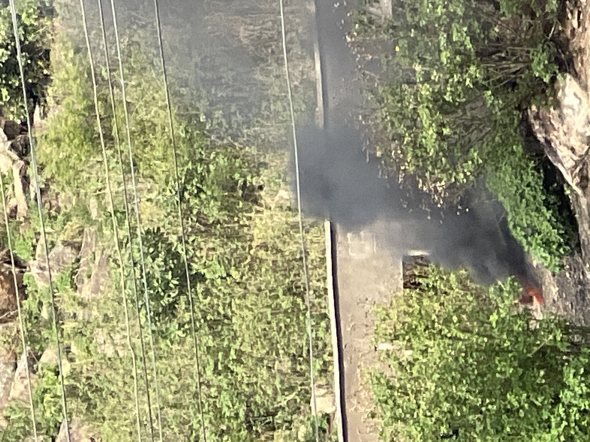 The image depicts a fire with black smoke billowing upwards, situated within a vegetated area next to a concrete structure. Overhead power lines are visible in the foreground. The location is Gura Top, Nigeria. There is no indication of people.