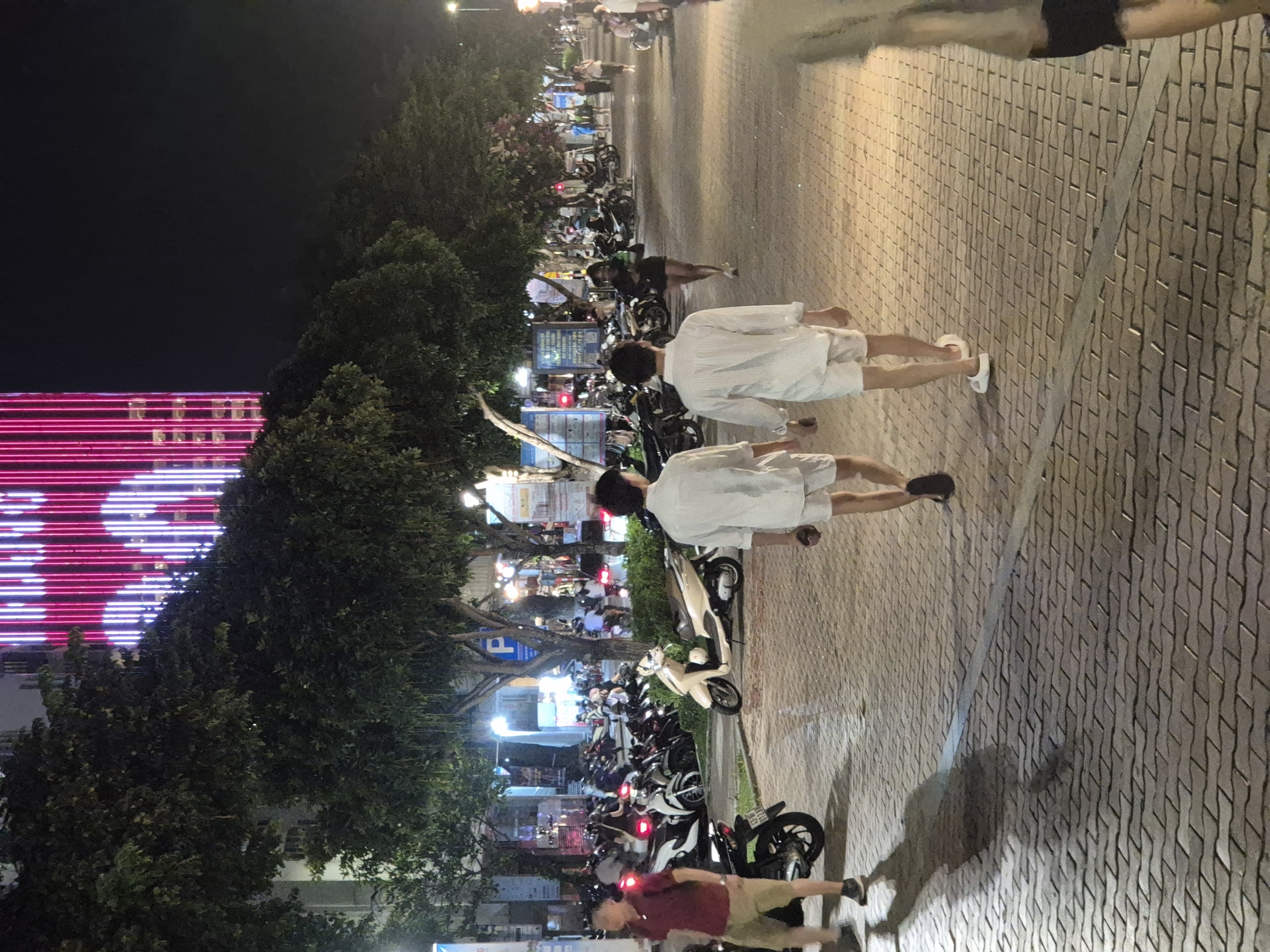 The image captures a night scene on a wide, paved sidewalk likely in the city of Đà Nẵng, Vietnam. The sidewalk is illuminated by streetlights and the vibrant neon lights of a building in the background, which features vertical pink and white stripes forming a heart shape. Trees with dense foliage line the sidewalk, casting shadows on the ground.
In the foreground, two young men are walking away from the camera. Both are wearing white, loose-fitting outfits consisting of button-down shirts and shorts. The man on the left is wearing white sandals, while the man on the right is wearing white slip-on shoes. To the left of these two men, a man in a maroon polo shirt and beige shorts is walking towards the camera.
Further down the sidewalk, a variety of parked motorcycles are visible, indicating a common mode of transportation in the area. Other pedestrians can be seen in the middle ground and background, some walking and others conversing. The overall atmosphere suggests a lively urban environment at night.