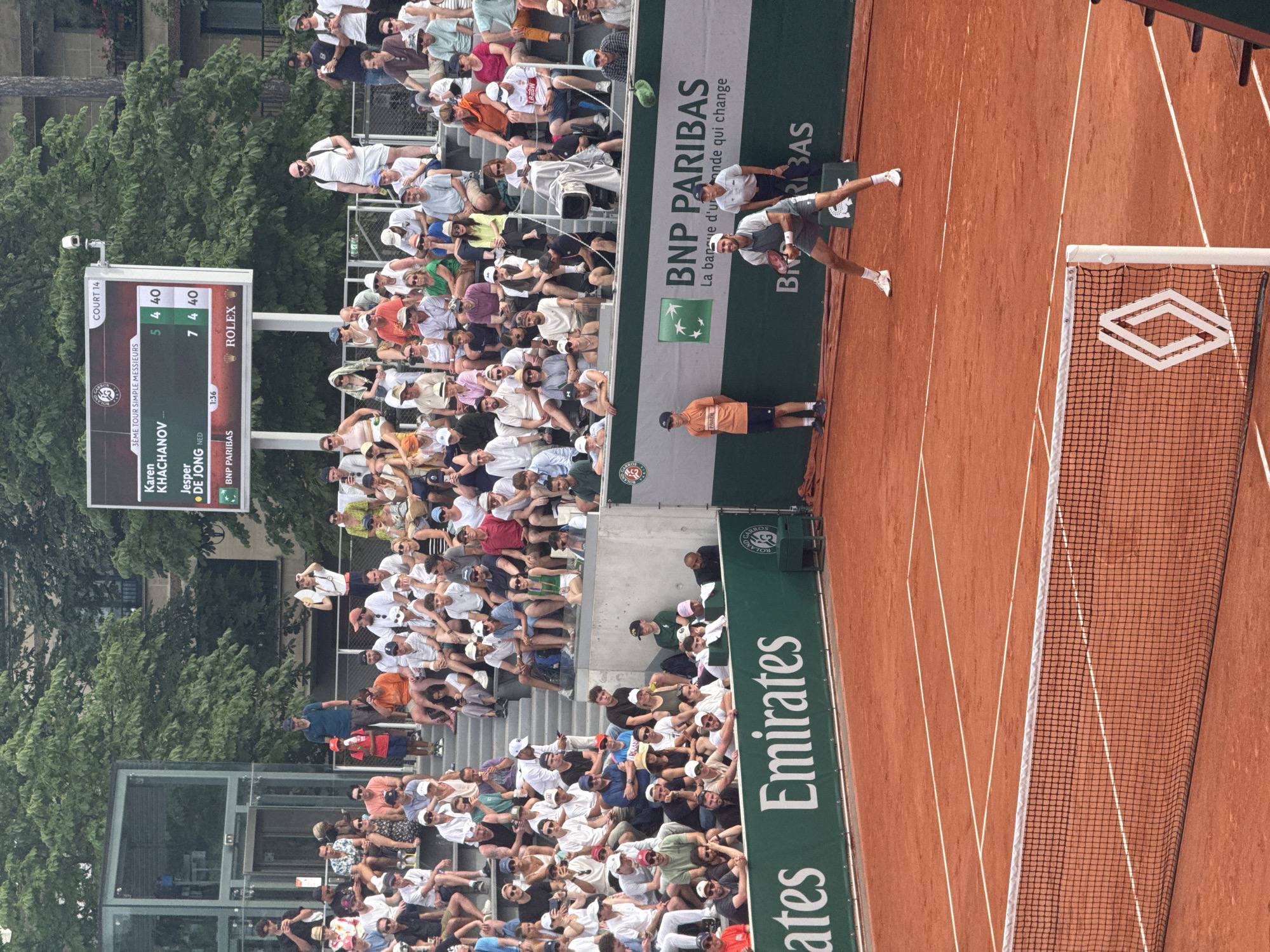 This image depicts a tennis match in progress at Roland Garros, Paris, France. The scoreboard indicates that it is a third-round singles match between Karen Khachanov and Jesper De Jong. Khachanov's score is 5-7, 4-4, while De Jong's score is 4-7, 4-4. A player is in motion on the clay court near the net. A linesman is visible on the court near an advertisement for BNP Paribas, one of the tournament's sponsors. The crowd fills the stands, and advertisements for Emirates and Rolex are also prominently displayed. Trees are visible behind the stadium seating.