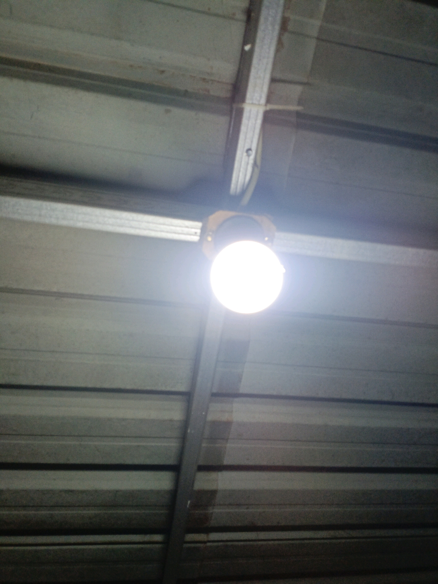 A bright, circular light bulb illuminates the underside of a corrugated metal roof. The bulb is encased in a yellowish fixture and is suspended by wires from a metal beam that runs horizontally across the frame. The corrugated metal panels are silver and appear to be part of a building's ceiling or roof structure. The lighting is overhead, casting a strong glow that causes some parts of the image to be overexposed. The environment suggests an interior space, possibly a workshop, warehouse, or unfinished building, with industrial-style construction. There are no people or specific details that would indicate a location within the city of Jenu, Indonesia.