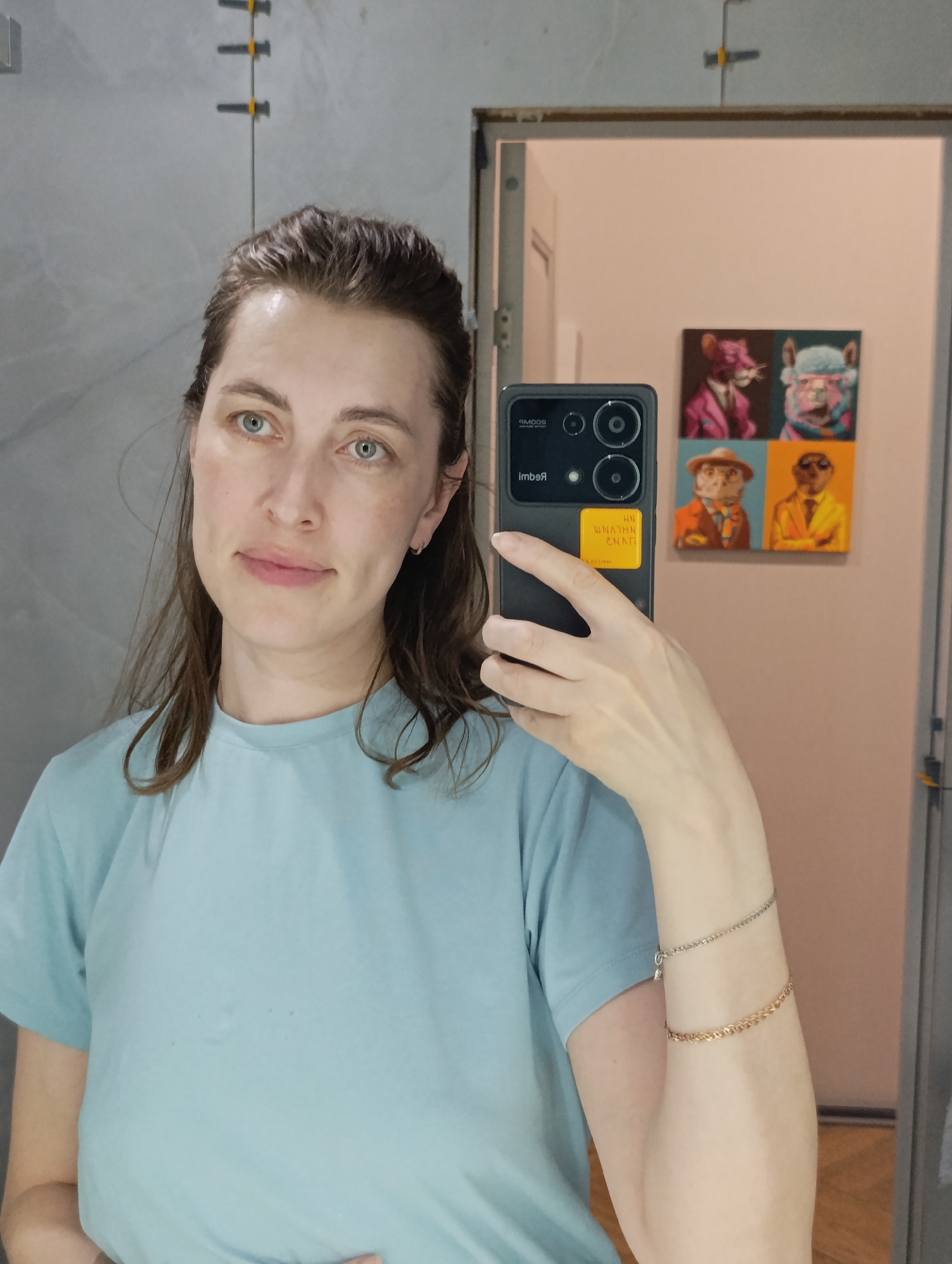 A woman is taking a selfie in a bathroom. She is wearing a light blue t-shirt and has two bracelets on her left arm. She is holding a black smartphone, and a yellow sticker is on the back of the phone. In the background, there is a mirror, a pink wall, and a painting of anthropomorphic animals. Yellow tile spacers are visible on the upper-left wall. The scene is indoors. No data indicates the location.