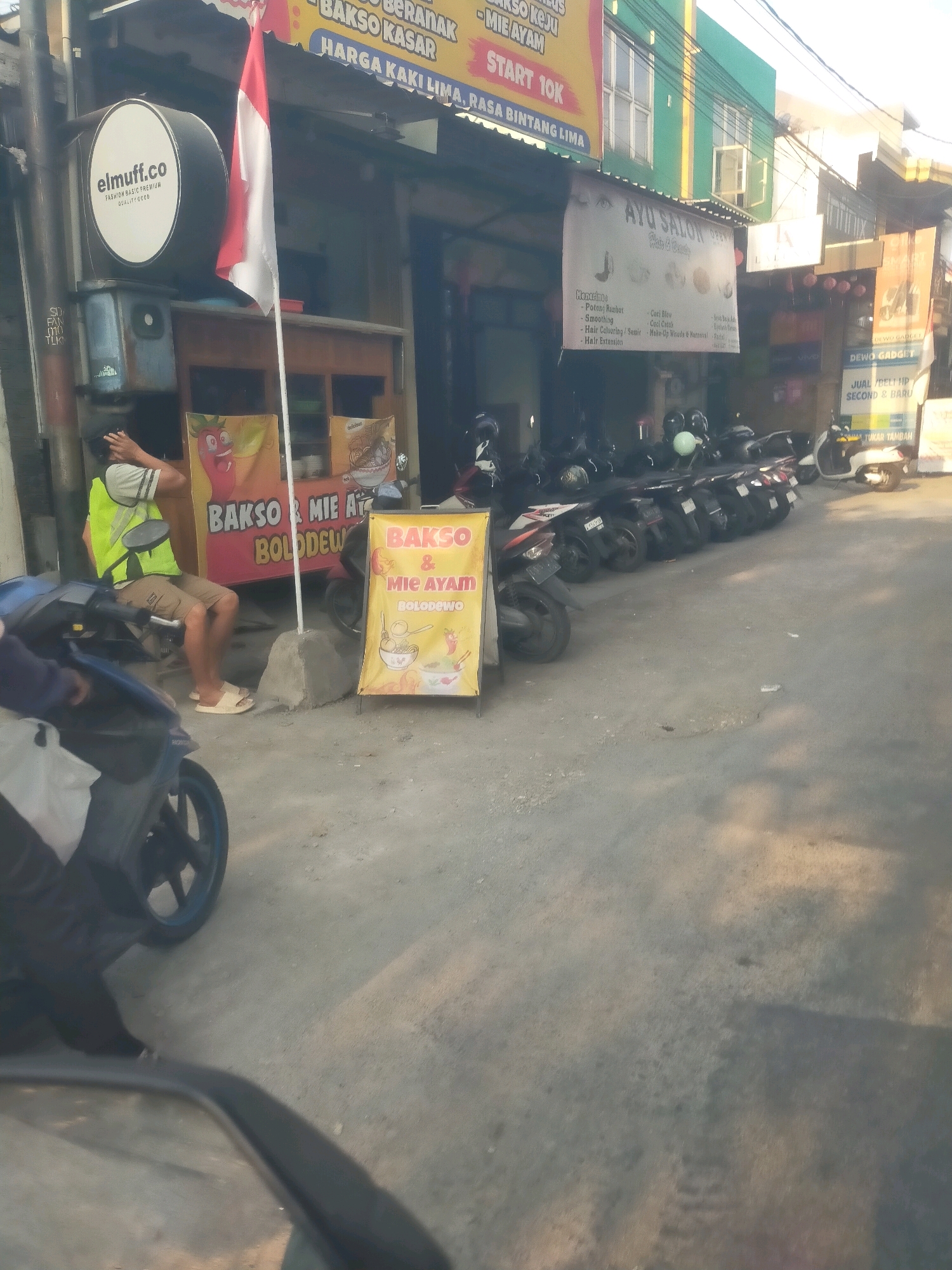 The image depicts a street scene in Sepande, Indonesia, likely in front of small businesses. In the foreground, a person wearing a bright yellow vest and light-colored shorts is seated on a motorcycle, holding their head. A red and white Indonesian flag is prominently displayed on a pole to the left of the frame. Several motorcycles are parked in a row along the street. A yellow banner advertises "BAKSO & MIE AYAM BOLODEWO," indicating a food stall selling meatballs and chicken noodles. Another banner nearby also advertises "BAKSO MI AYAM BOLODEWO". Further down the street, there are signs for businesses including "ELMUFF.CO" and "AYU SALON". The overall environment appears to be a busy urban street with shops and commercial activity.