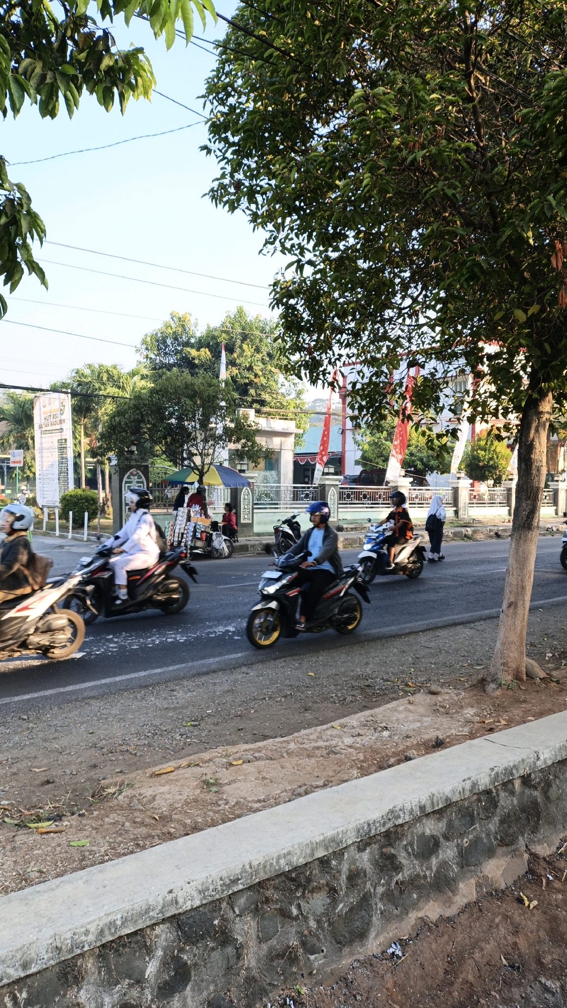 The image depicts a street scene in Kuwasen, Indonesia, characterized by the presence of several individuals riding motorcycles. The foreground is dominated by a concrete barrier with a stone foundation, bordering a dusty roadside. Beyond this, a paved road accommodates a moderate flow of traffic, primarily motorcycles. Several riders are visible, some wearing helmets, and at least one is in traditional Islamic attire. In the background, buildings with awnings, flags, and trees suggest an urban or semi-urban environment. A billboard with text in Indonesian is also present. The overall lighting indicates it is daytime, likely morning or late afternoon, given the long shadows and warm sunlight. The atmosphere appears to be that of a typical day on a busy street in Indonesia.