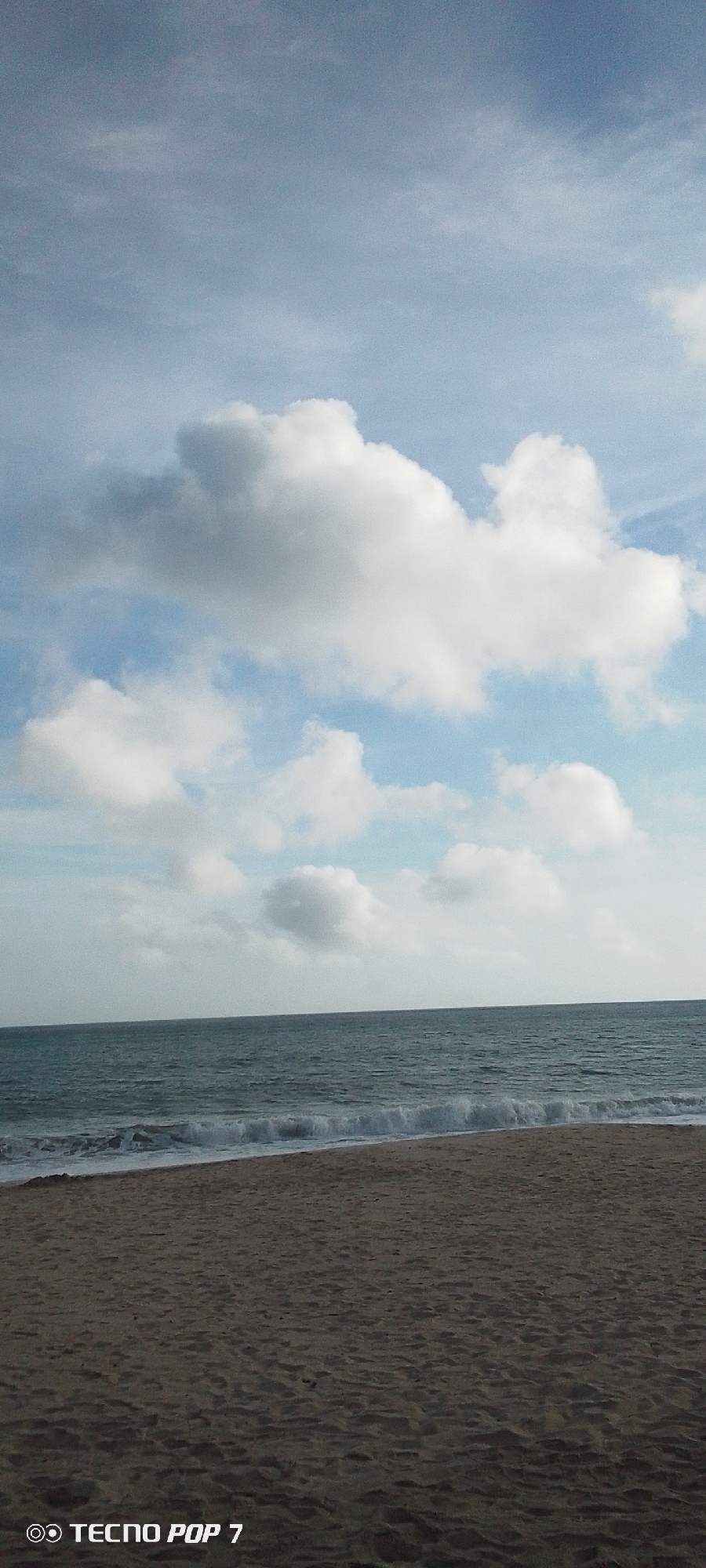 The image shows a beach scene in Pedro González, Venezuela. The foreground features a sandy beach with a dark tan color. The midground contains the ocean with small waves breaking on the shore. The background depicts a sky with scattered, puffy white clouds and patches of light blue. No people or objects are visible on the beach. The text "TECNO POP 7" appears in the lower left corner of the image.