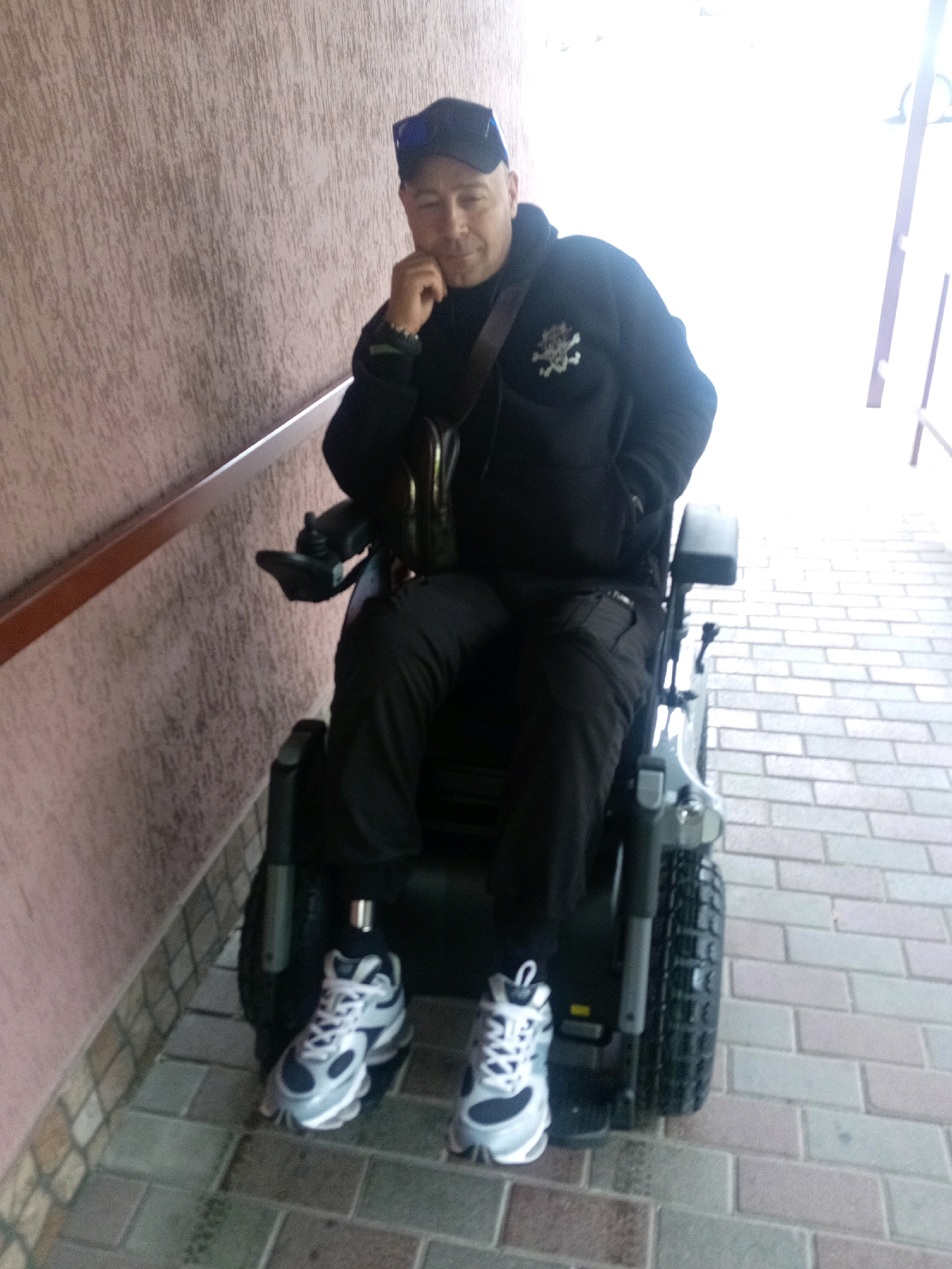 A man wearing a black hoodie, black baseball cap, and black pants is sitting in a motorized wheelchair. He has a dark-colored skull and crossbones emblem on the left side of his hoodie. He is resting his chin on his hand and looking towards the camera. He is wearing white and grey sneakers. The wheelchair is grey and black with a black tire. The man is positioned on a paved walkway with a textured, pinkish-brown wall on the left side. The walkway is made of interlocking brick pavers. The scene appears to be outdoors, possibly in front of a building. The lighting suggests it is daytime.