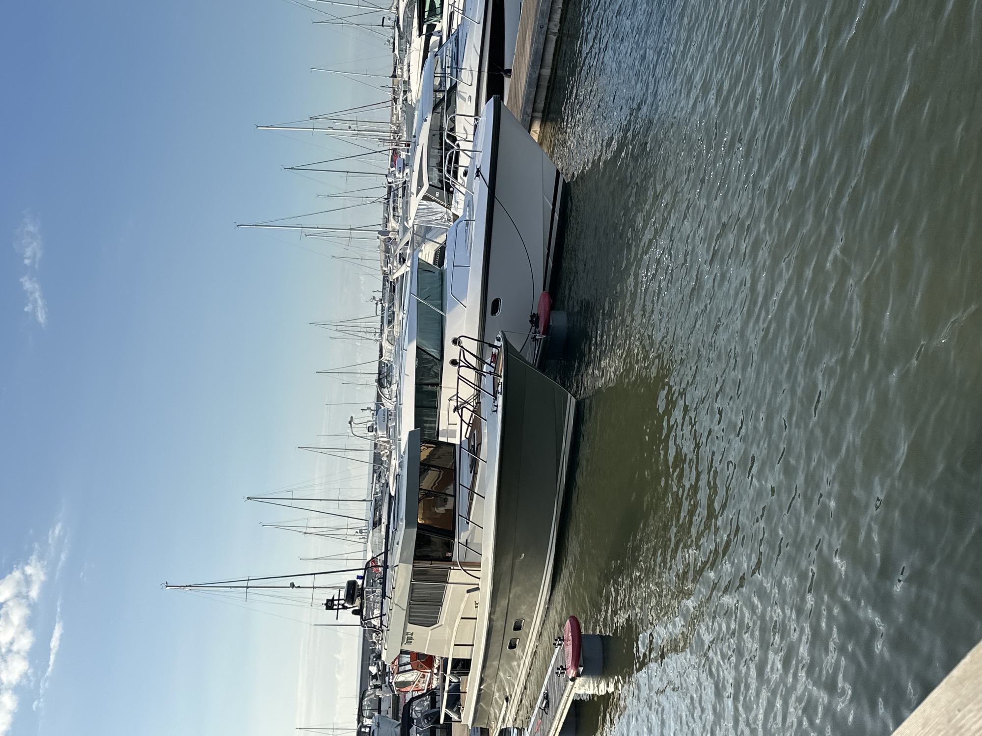 The image shows a marina scene in Tallinn, Estonia, with several boats docked. In the foreground, a grey motorboat is prominently featured, with its name "Targa 32" visible on the side. To its right, a white motorboat is partially visible. In the background, numerous sailboats with tall masts are lined up, creating a picturesque harbor view. The water is calm and reflects the blue sky and the boats. The sky is clear with a few wispy clouds. The scene appears peaceful and sunny, suggesting a pleasant day at the marina.