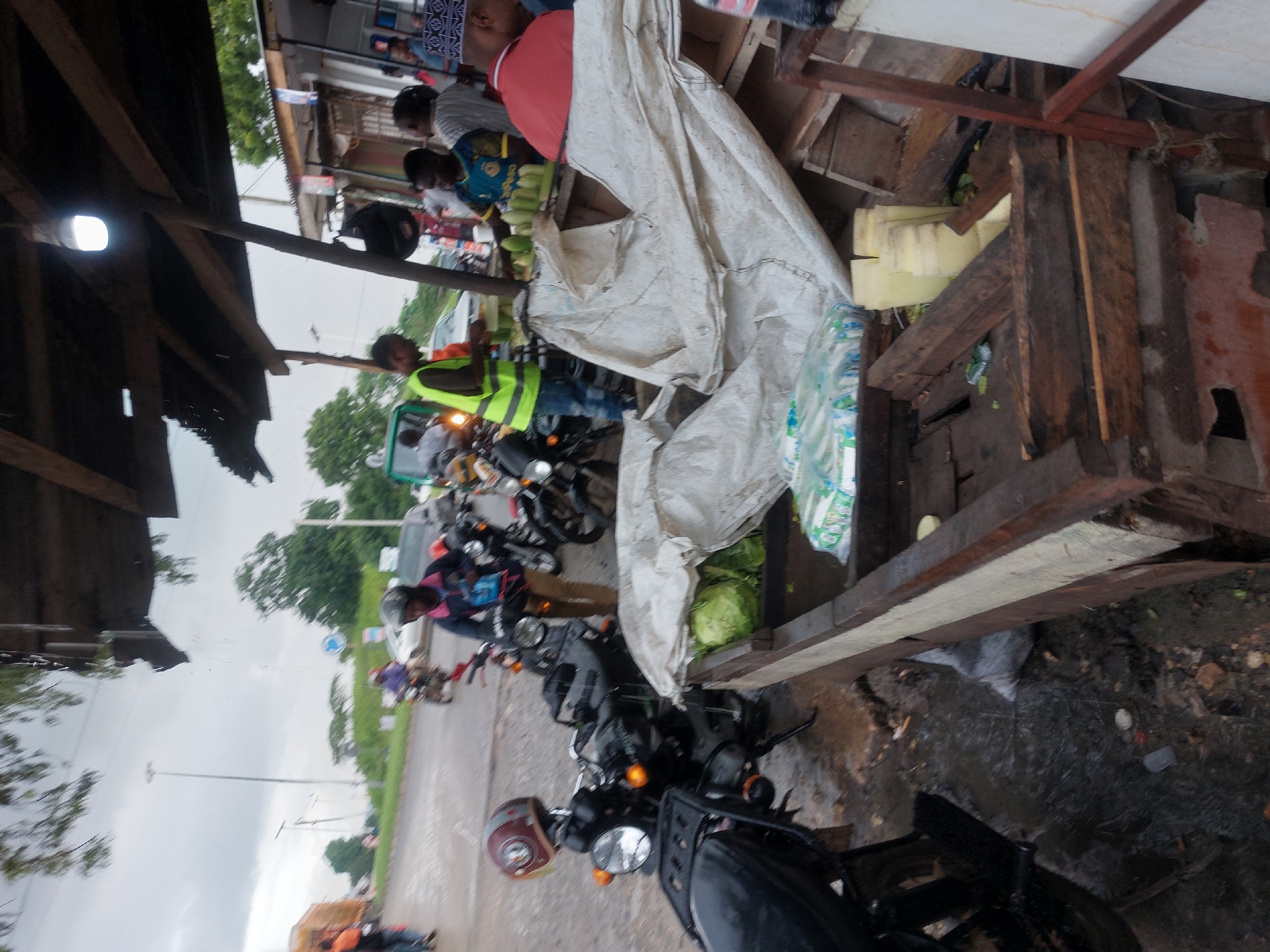 The scene depicts a roadside vendor stall in an urban setting, potentially in Dar es-Salaam, Tanzania, based on the visual characteristics. Several people are visible, some standing near the stall and others sitting on motorcycles parked along the road. The vendor stall has a wooden structure and is covered with a white cloth, with some produce visible beneath. The road appears wet, possibly from recent rain. A row of motorcycles is parked adjacent to the stall, with several individuals either on or near the bikes. The overall environment suggests a busy, informal market or transportation hub.