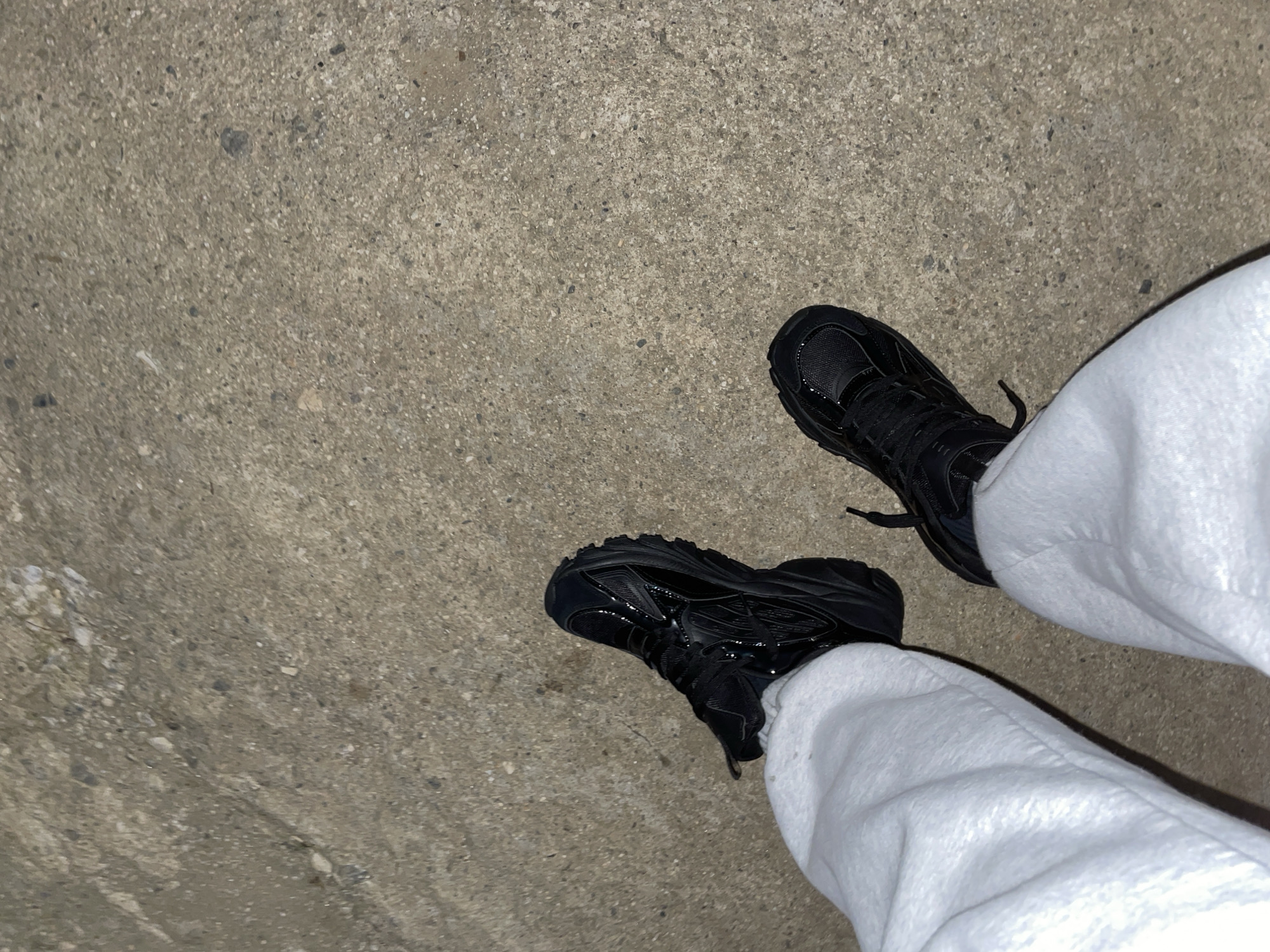 The image shows a view of a pair of black sneakers worn with white sweatpants. The footwear is placed on a concrete surface. The scene appears to be photographed outdoors, likely on a sidewalk or similar paved area. There are no people or specific actions visible other than the wearing of the shoes. The background is a grey concrete surface with a somewhat rough texture. There is no indication in the image that it is related to or located in Zlatograd, Bulgaria.