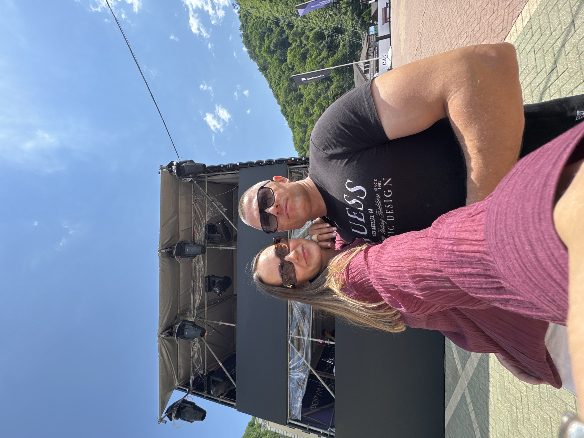 This is a selfie of a couple outdoors during the day. The man, on the left, wears a black t-shirt with white text that says "GUESS LOS ANGELES, CA SINCE 1981 Lasting Tradition Music DESIGN". He is wearing sunglasses. The woman, on the right, wears a pink, textured, long-sleeved top and sunglasses. They are standing in front of a large black structure, possibly a stage or display for an event, with scaffolding and lighting equipment visible. In the background, a lush green, tree-covered mountainside rises under a bright blue sky with scattered white clouds. Flags with Cyrillic text are visible on the mountainside. The ground is paved with light-colored bricks. The scene appears to be set in a tourist area, possibly a resort, given the mountains and paved walkway. The overall mood is casual and bright, suggesting a pleasant outdoor activity. The text visible on the structure reads "ФОРУМ НА ИЖИЛ ССТІ" and "CA".