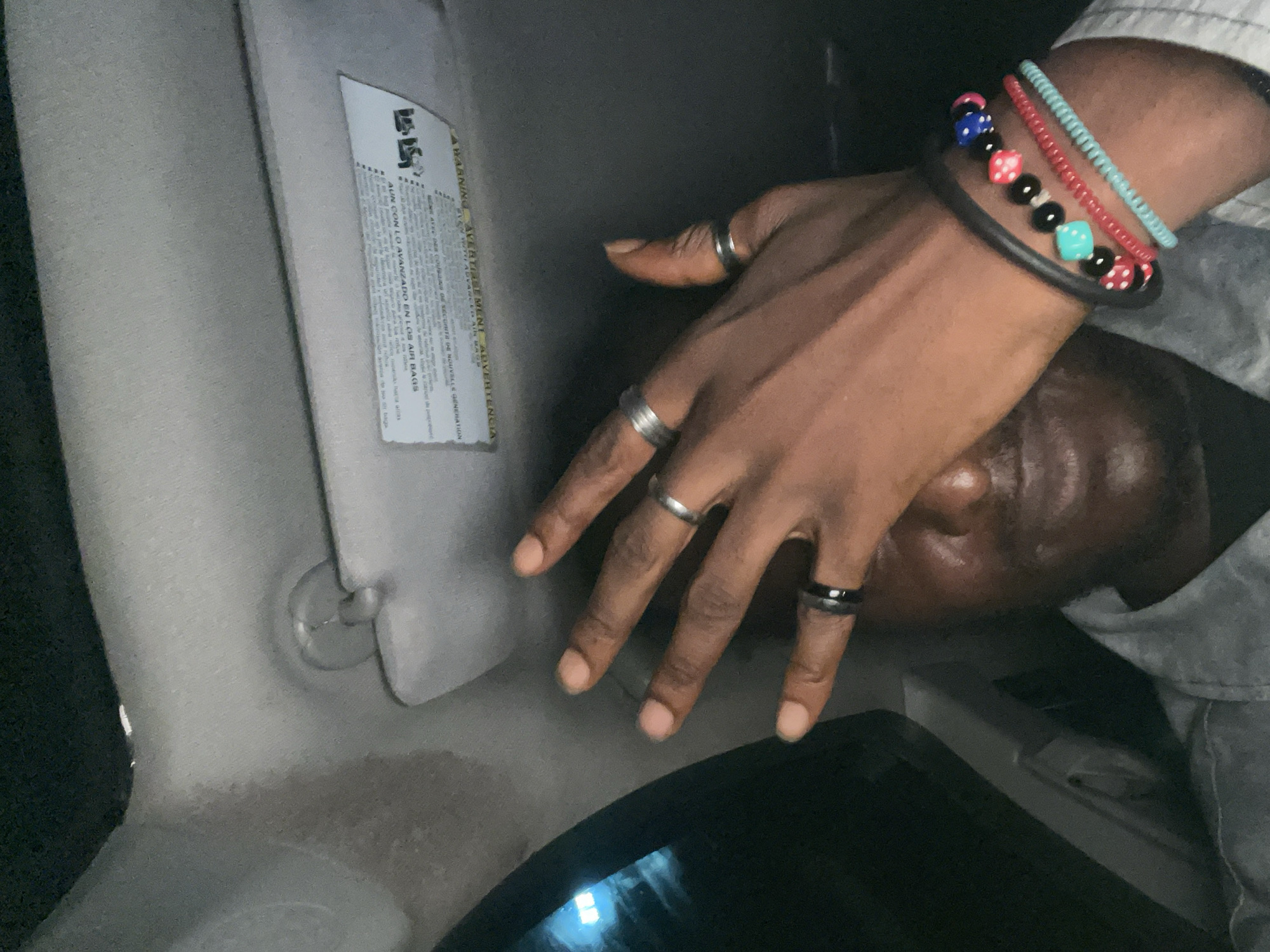 The image shows a close-up of a person's right hand and wrist, partially obscuring their face. The hand has several rings on it, including a silver band on the ring finger, a wider silver band on the middle finger, and a black band on the index finger. The wrist is adorned with multiple bracelets. One is a black leather band, and stacked on top of it are beaded bracelets, featuring black beads, red beads with white dots, blue beads with white dots, and a light blue bead with white dots. The person is wearing a light-colored collared shirt. The background appears to be the interior of a car, with a visible sun visor overhead that has text printed on it. The lighting suggests it might be nighttime or a dimly lit environment. There are no discernable landmarks that place the image specifically in Abuja, Nigeria, or any other city.
