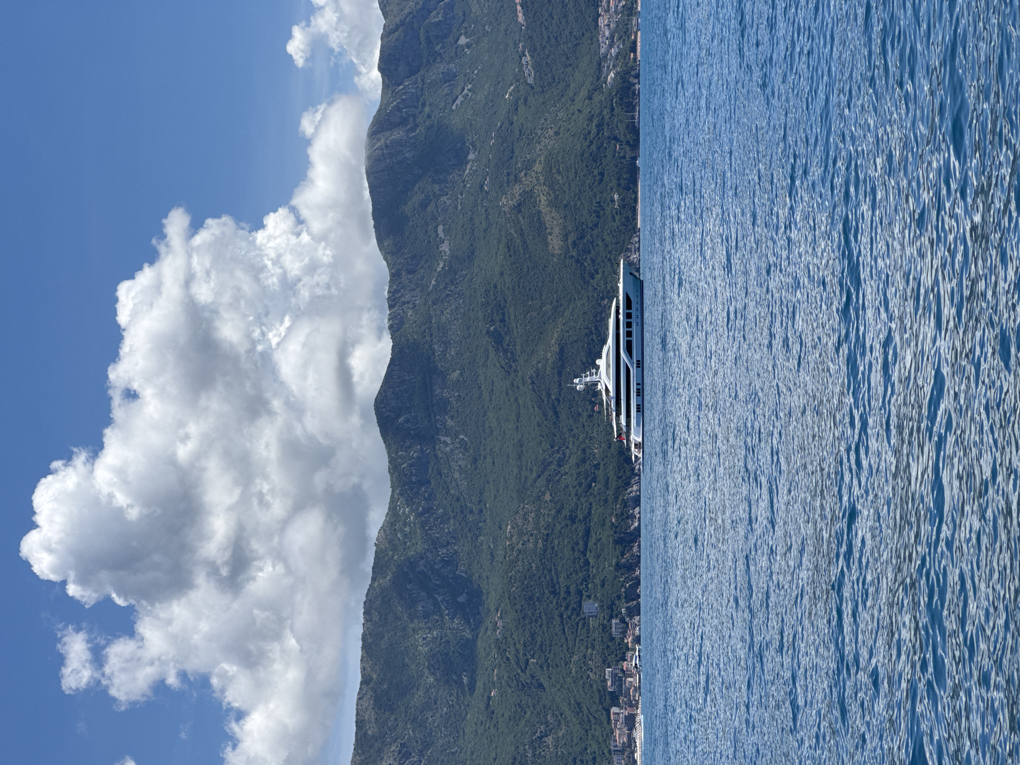 The image, best viewed horizontally, captures a serene coastal landscape under a bright, clear blue sky. Dominating the foreground and extending to the right is a wide expanse of deep blue water, showing subtle ripples and reflections. A large, white, multi-deck yacht is visible on the water in the middle-ground, positioned closer to the mountainous coastline, its pristine exterior contrasting with the blue sea.
In the background, a majestic, dark green mountain range stretches across the horizon. These mountains are densely covered in vegetation, indicating a lush environment, and they appear to rise steeply from the coastline. At the base of these mountains, nestled along the shore, a collection of buildings suggests a bustling town or city, characteristic of coastal settlements.
The sky above features large, fluffy white cumulus clouds, concentrated on the left side of the frame, adding depth and visual interest to the expansive blue. The scene is brightly lit, indicating a sunny, clear day, likely around midday, given the light quality and the shadows. The overall impression is one of natural beauty and tranquility, typical of a leisure setting in Budva, Montenegro. No text is visible in the image.