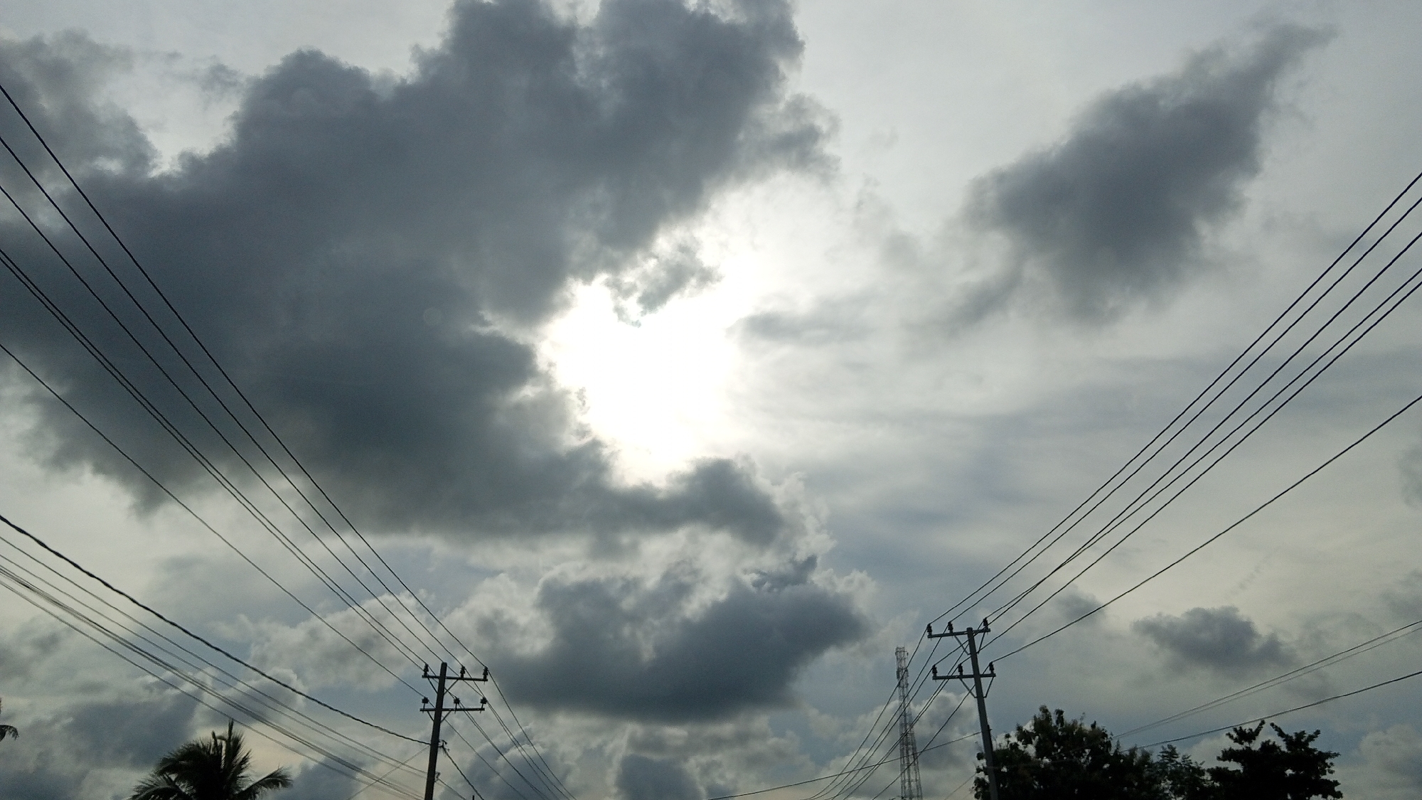The image captures a cloudy sky over Payameuneng, Indonesia, with a bright, diffused sun peaking through a gap in the thick, dark clouds. Multiple power lines, supported by utility poles, crisscross the frame, drawing the eye upwards towards the dramatic sky. In the lower portion of the image, silhouetted palm trees and other foliage are visible, suggesting a tropical or sub-tropical environment. The overall mood is somber and atmospheric, indicative of an impending storm or the aftermath of one. There are no people or animals visible in the frame.