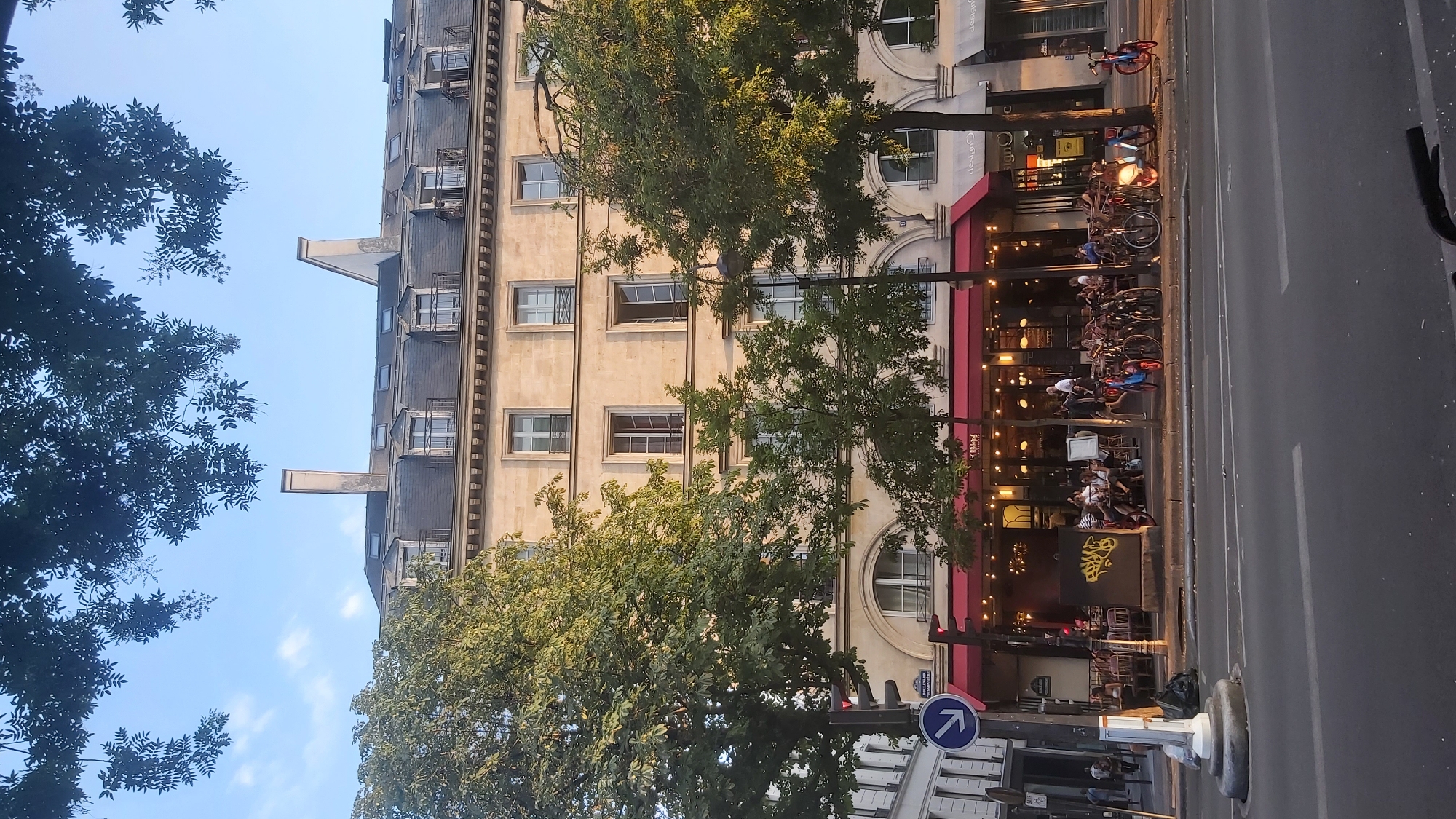 The image captures a street scene in Paris, France, under a clear blue sky with scattered clouds. A multi-story building with ornate architecture dominates the background. The building has a mansard roof with dormer windows and a light-colored stone facade. In the foreground, a tree-lined sidewalk fronts a bustling outdoor cafe with red awnings. Numerous patrons are seated at tables, engaged in conversation and dining. A row of bicycles is parked along the sidewalk, suggesting a common mode of transportation in the area. Traffic lights and street signs are visible, indicating an urban setting. The lighting suggests it is either early morning or late afternoon, with the sun casting long shadows. The overall atmosphere is one of leisurely urban life in Paris.
