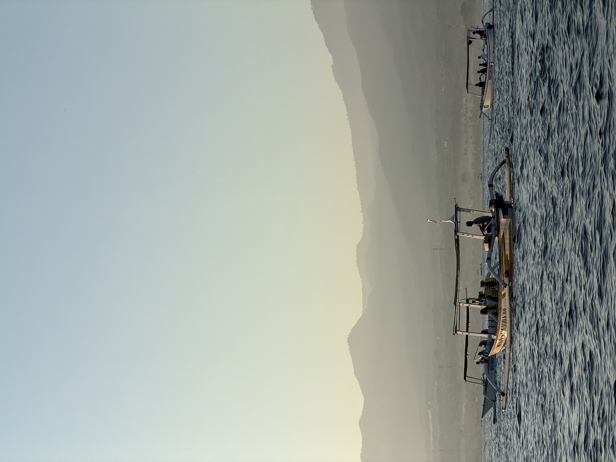 In the city of Buleleng, Indonesia, on a body of water with moderately rippled dark blue waves, three outrigger boats are visible. The boat in the foreground is yellow and white with an overhead canopy, carrying at least seven people wearing life vests. A person is steering from the back. The boat in the middle ground is white with a similar canopy, carrying at least four people, two of whom appear to be wearing life vests. The boat on the far right is also white with a canopy and carries at least four people. The background features hazy, layered mountains under a clear, pale blue sky that transitions to a pale yellow near the horizon. The overall lighting suggests it is either early morning or late afternoon.