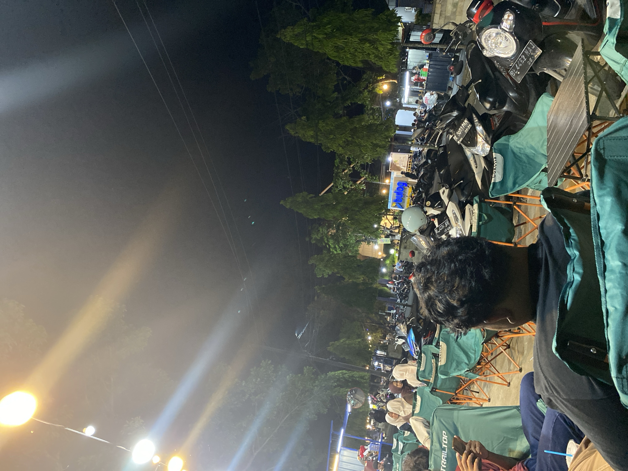The image depicts an outdoor gathering at night, likely a food court or a casual seating area, in Lhokseumawe, Indonesia. A row of green camping chairs, some with people seated in them, is arranged in the foreground. Several motorcycles are parked in the background, indicating this is a public space where people arrive by vehicle. Strings of warm-toned lights are hung overhead, illuminating the scene. In the distance, there are more people gathered, suggesting a lively atmosphere. The presence of trees and a dark sky point to an urban or semi-urban environment. The overall impression is of a relaxed, communal setting for dining or socializing in the evening.