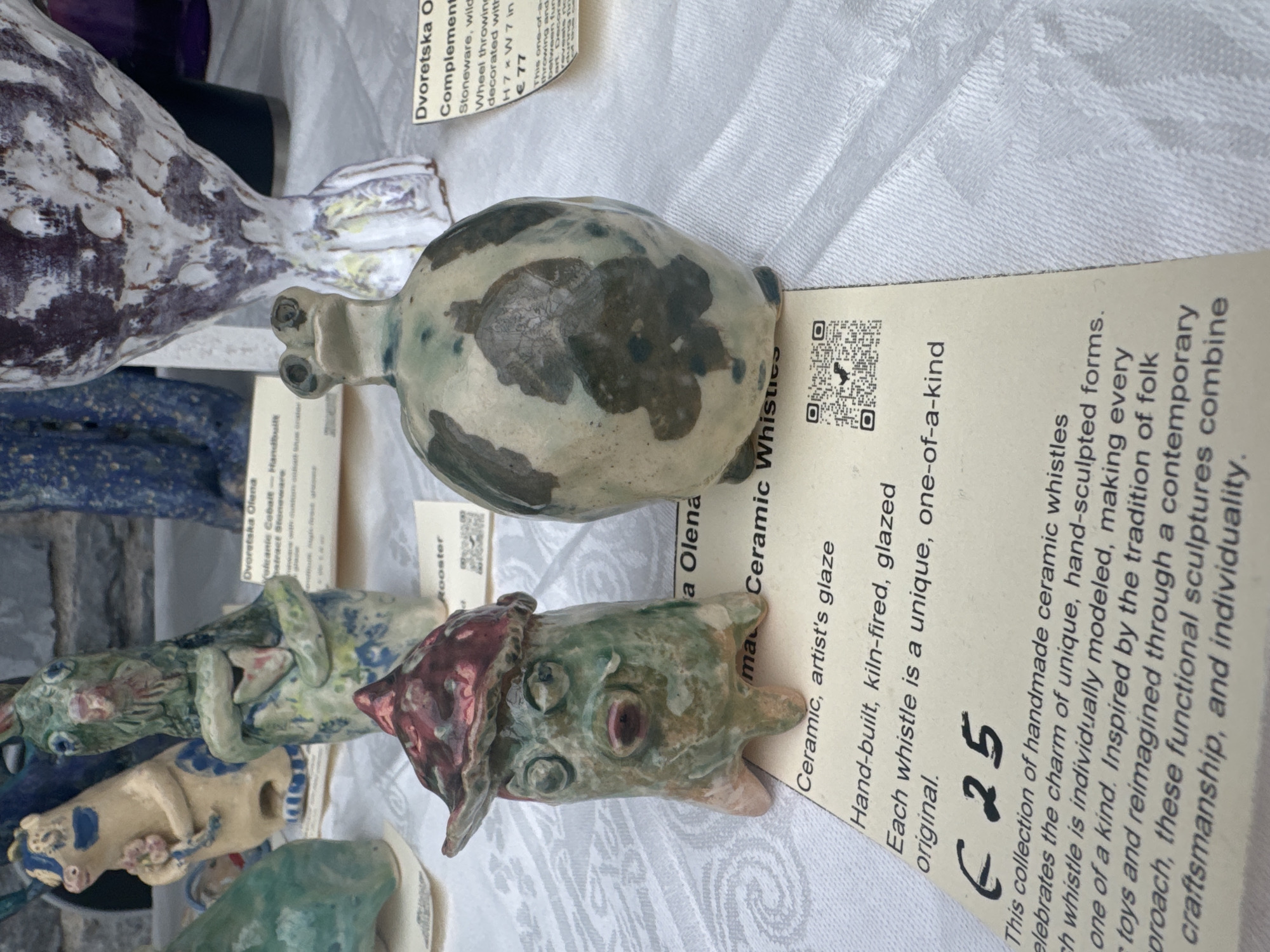 The image displays a selection of handmade ceramic whistles displayed on a white tablecloth. In the foreground, a small, green, anthropomorphic figure with a red hat is visible, standing on four small legs. To its right is a larger, bulbous ceramic object with dark green markings. Both are positioned on paper tags with handwritten and printed text. One tag, partially obscured, mentions "Dvoretska Olena," "Rooster," and "Ceramic, artist's glaze." Another tag details "Hand-built, kiln-fired, glazed," and states "Each whistle is a unique, one-of-a-kind original." The price "€ 25" is clearly marked. Further back, other ceramic figures and objects, some featuring blue and white patterns, are visible. The background suggests an outdoor market or craft fair setting. The overall impression is a display of artisanal pottery for sale. The text on the tags, particularly the price, implies this is a retail environment. While the image itself does not contain direct evidence of Kilkenny, Ireland, the presence of such a craft display is common in artisan markets in cities like Kilkenny, known for its cultural heritage and craft scene.