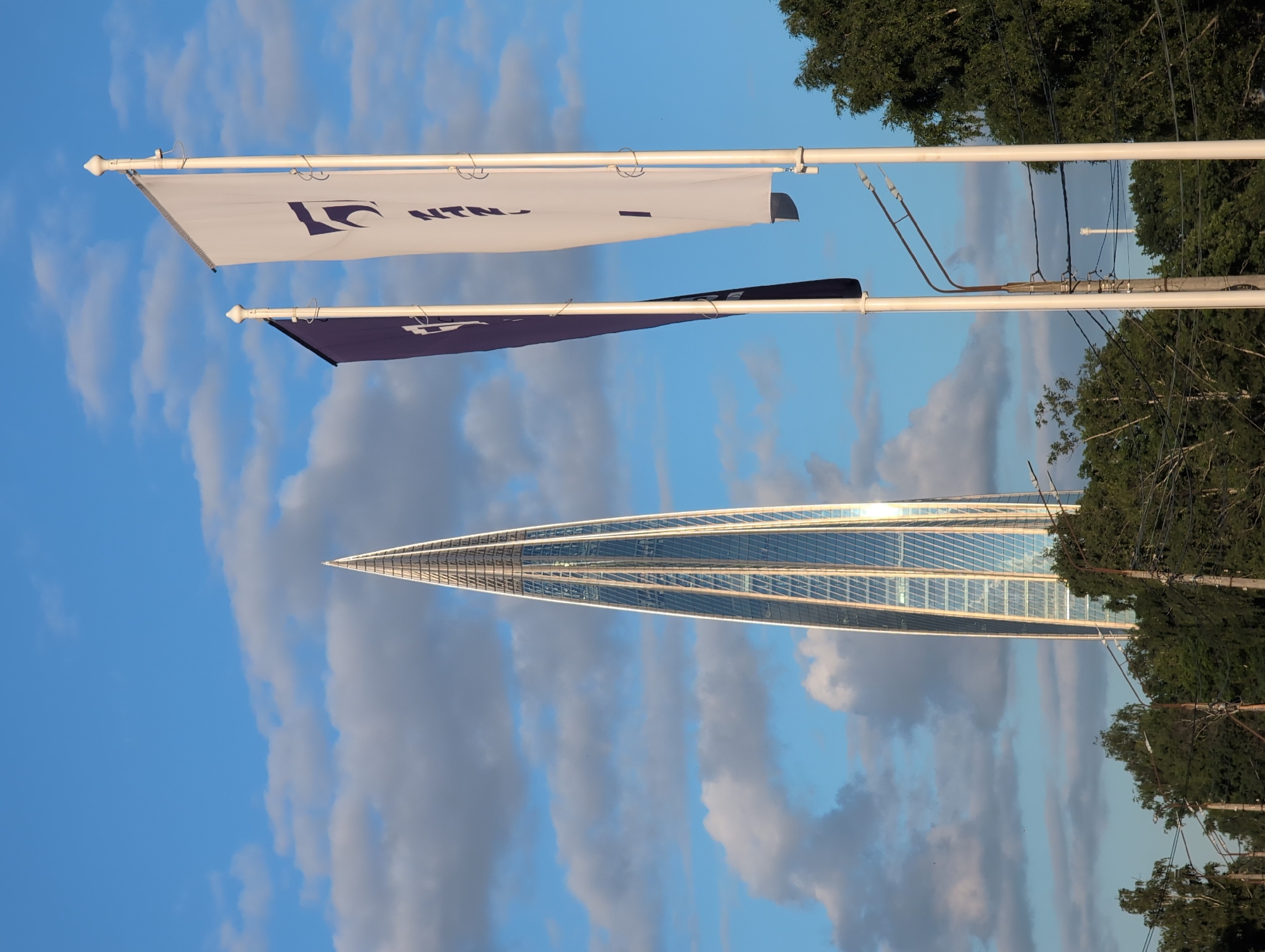 The image captures a daytime scene in Saint Petersburg, Russia, under a sky filled with scattered clouds. The primary subject is a strikingly modern, twisting glass skyscraper, its reflective facade catching the light. In the foreground, two flags hang from poles. The upper flag is white with a dark blue logo and text that appears to read "IC NTN." Below it, a dark blue flag is also visible, though its details are less clear. Trees and power lines are visible to the right of the skyscraper, suggesting an urban or suburban environment. The lighting indicates it is likely late afternoon or early evening, given the warm tones on the skyscraper. The overall mood is one of architectural grandeur set against a natural backdrop of sky and foliage.