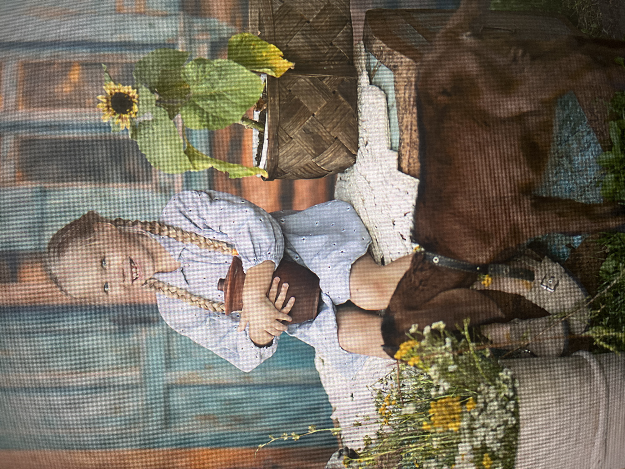 A young Caucasian girl with blonde braided hair and gaps in her front teeth is sitting on a wooden surface in front of a weathered blue wooden door. She is wearing a light blue dress with a small white pattern and is holding a brown clay pot. To her right, a sunflower plant is in a woven basket. In the foreground, a brown goat is eating from a pot of yellow and white flowers. The setting appears to be outdoors, possibly a rustic courtyard, and given the context of Izhevsk, Russia, it could be a rural or semi-rural area within or on the outskirts of the city.