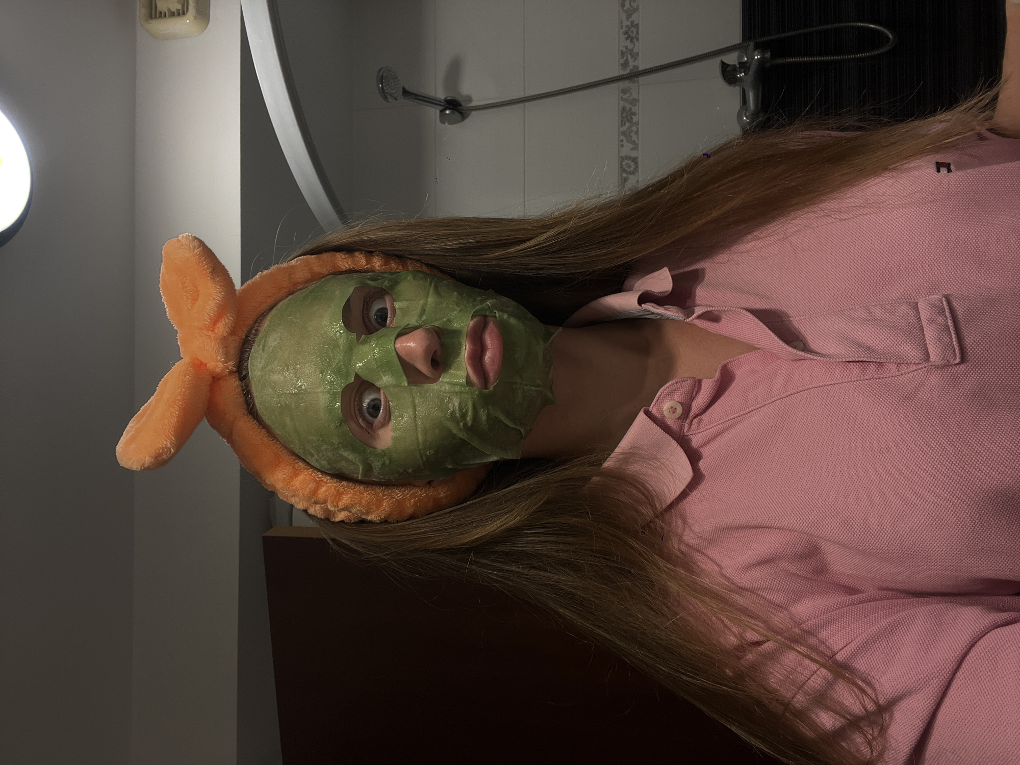 The image captures a close-up of a person engaged in a self-care routine, oriented on their side. The subject has long, flowing light brown hair and is wearing a green sheet face mask that covers most of their face, with cut-outs revealing wide-open blue eyes, their nose, and slightly parted pink lips. An orange, plush headband with two ear-like structures is worn on their head, keeping the hair away from their face.
The person is dressed in a pink short-sleeved polo shirt, notable for its textured fabric and visible collar and buttons. The background suggests an indoor setting, likely a bathroom, with white tiled walls. A chrome showerhead with a flexible hose is mounted on the wall above the person's head. Further up on the wall, a small, light-colored object, possibly a soap dish, is visible. To the left, a bright, circular artificial light source illuminates the scene, and a curved, reflective surface, possibly a mirror, is partially visible.
The activity depicted is the application of a face mask for skincare. The eyes are looking directly at the camera, suggesting an intentional photo. The artificial lighting indicates that the photo was likely taken indoors, possibly in the evening or at night. No visible text is present within the image.