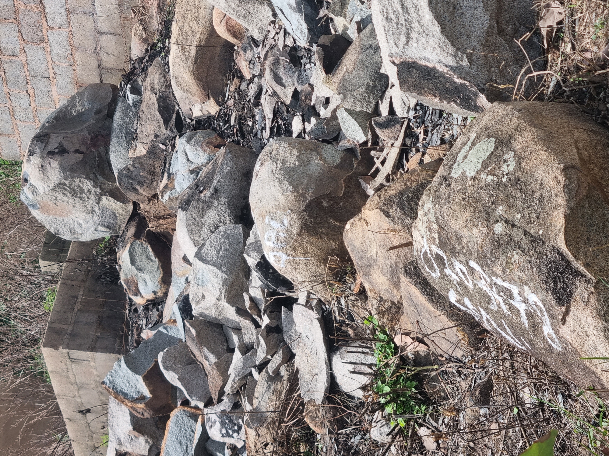 The image depicts a pile of large rocks and boulders in a natural setting. Some of the rocks have white markings on them, possibly graffiti or numbering. In the background, there is a concrete structure and a brick wall, suggesting the presence of human construction nearby. The ground is covered with smaller stones, dry vegetation, and some patches of green grass. The overall scene appears to be an outdoor area, possibly a construction site or a quarry, located in the city of Jos, Nigeria.