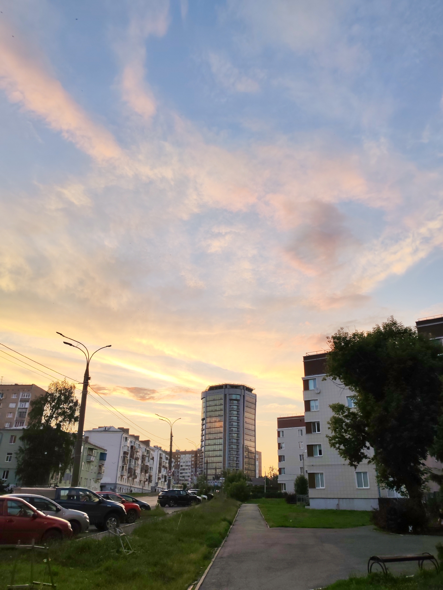 This is a vertical, eye-level, outdoor, full shot of a city street scene in Izhevsk, Russia during sunset. The sky is filled with wispy clouds tinged with shades of pink, orange, and yellow, contrasting with the pale blue sky above. In the mid-ground, a tall, modern, cylindrical glass building with reflective windows stands out, catching the sunset light. To the right of this building is a multi-story apartment complex with a light-colored facade and visible windows. In the foreground, on the left side of the image, is a row of parked cars, including a red sedan, a silver sedan, a dark SUV, and another dark car. To the right of the cars is a grassy area and a paved walkway. Several streetlights and utility poles with wires are visible. Trees and other buildings are scattered throughout the scene. The overall impression is a quiet urban evening.