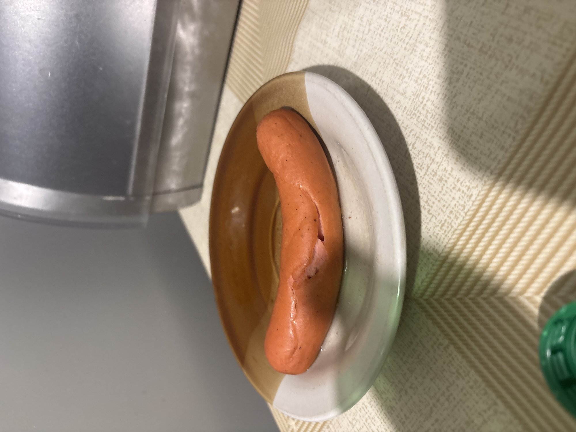A cooked sausage lies on a beige and white plate. The plate sits on a beige and white textured surface, likely a tablecloth. Part of a metallic object is visible to the left of the plate. The location and city of Yerevan, Armenia, cannot be determined from the visual information provided.