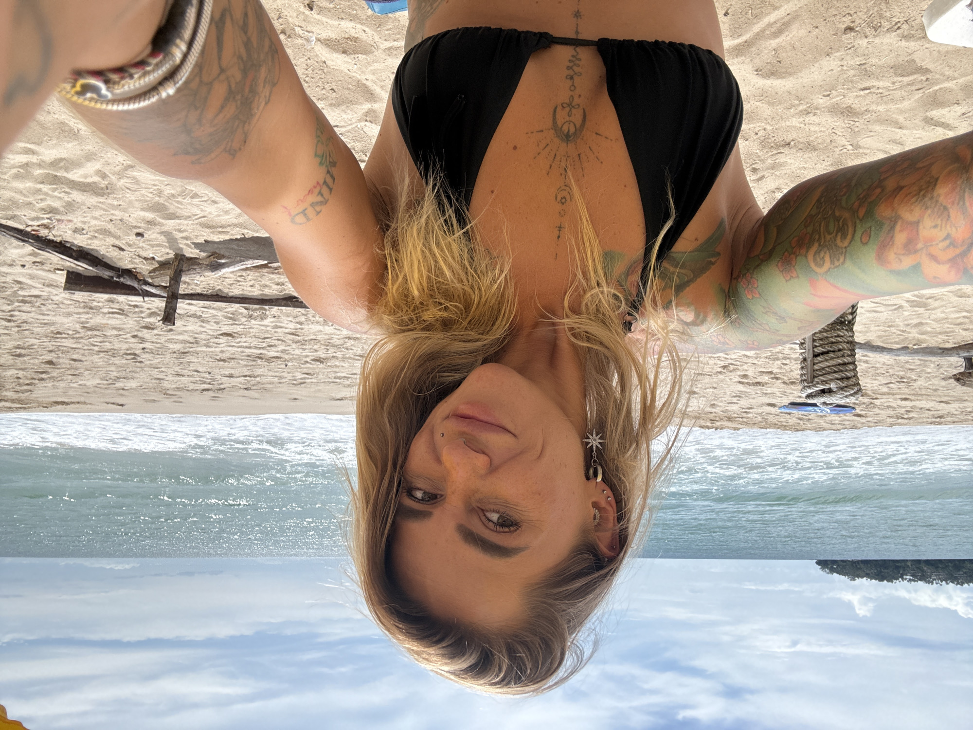 This is an upside-down, close-up selfie of a woman on a beach. She has blonde hair and is wearing a black bikini top. She has tattoos on her arms and chest. Her left arm, which is higher in the frame, has a tattoo with the word "MIND" and a lotus flower. Her chest tattoo appears to be a complex geometric design with celestial elements. She is wearing earrings, including a star-shaped one in her left ear. The background shows a sandy beach, the ocean with waves, and a cloudy sky. The setting is likely tropical, given the location context of Choeng Thale, Thailand. The lighting suggests it is daytime. The woman's expression is a slight smile.