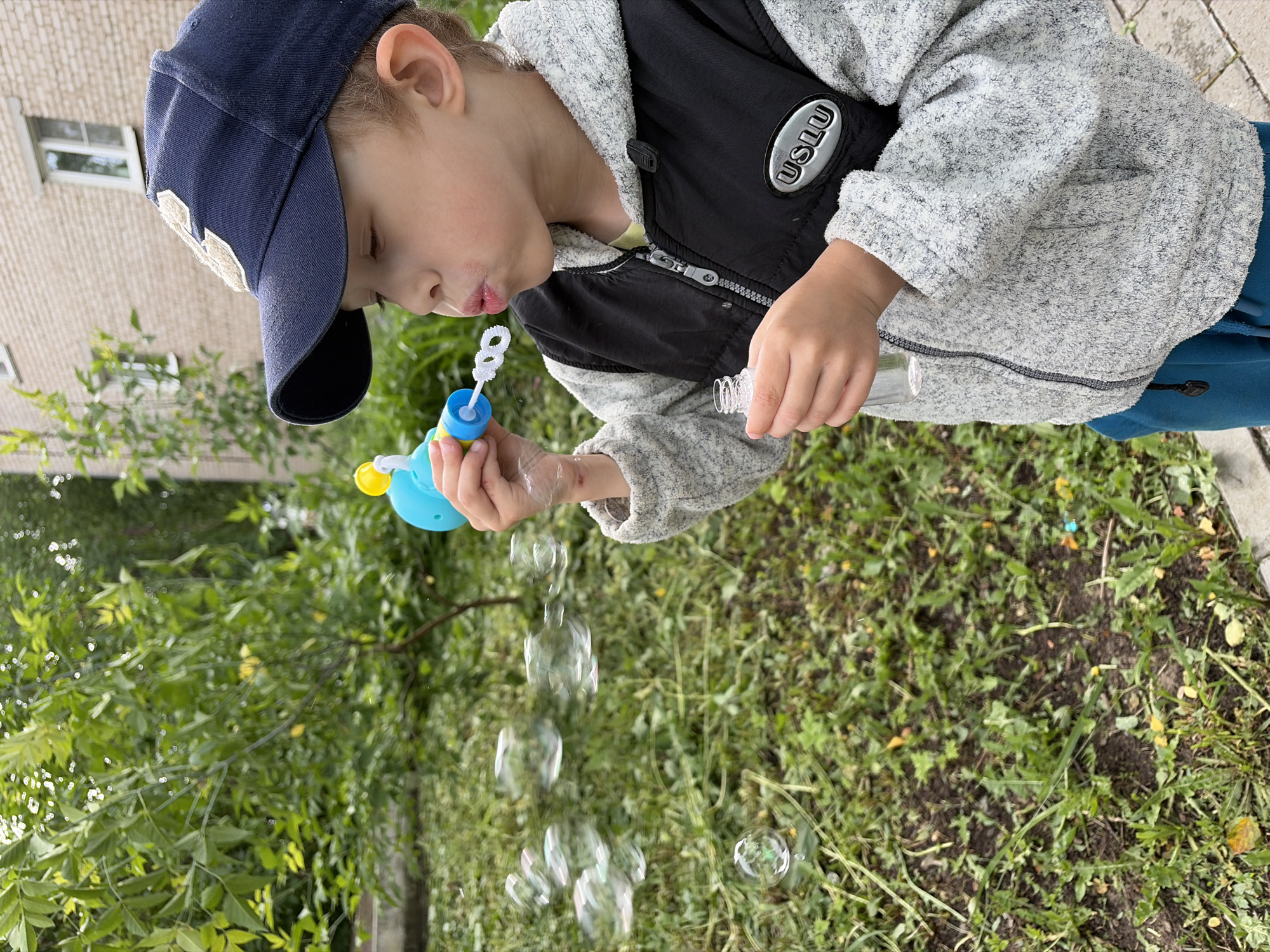 A young child is shown outdoors, actively blowing soap bubbles. The image is captured in a vertical orientation but displayed rotated clockwise, placing the ground to the right and a building to the left.
The child, likely a boy, is wearing a dark blue baseball cap with a white embroidered emblem, a light grey textured hooded jacket, and a black puffer vest over a lighter shirt. Blue pants are also visible. He holds a blue and yellow bubble wand to his mouth with his left hand, appearing to blow bubbles, while his right hand holds a small clear plastic bottle of bubble solution. Several shimmering, transparent soap bubbles are visible in mid-air, floating downwards from the wand. The child's expression conveys concentration and engagement in the activity.
The setting is an outdoor space, possibly a yard or park, with lush green foliage, including bushes and grass, forming the background and foreground. In the upper left corner, a light-colored brick building with a white-framed window is partially visible. The ground cover consists of grass and some exposed soil.
The scene is illuminated by bright daylight, suggesting fair weather. A notable detail is the word "USLU" displayed on a silver patch affixed to the child's black vest. The event takes place in Chaikovsky, Russia.